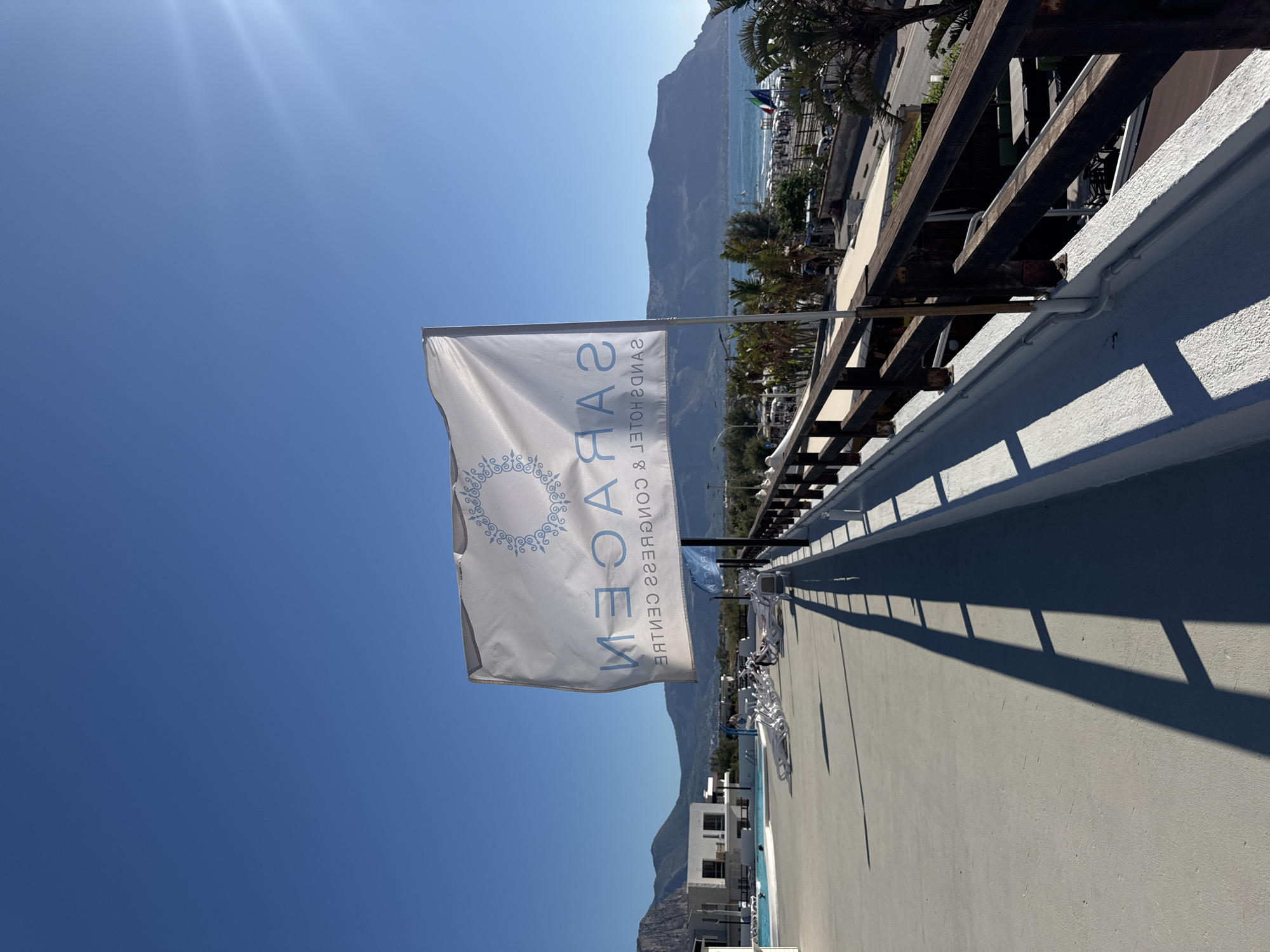 A white flag with a blue circular emblem and blue text is prominent in the foreground. The text on the flag reads "SARACEN SANDS HOTEL & CONGRESS CENTRE", however, it appears to be reversed. The flag is attached to a pole and is waving in the wind. The flag is casting a long shadow on a light-colored concrete surface.
The background reveals a sunlit scene under a clear blue sky with the sun's rays visible in the upper right corner. Beyond the concrete area, there is a dark wooden railing. Further in the distance, there is a body of water, which appears to be the sea, with a beach lined with umbrellas and people. Behind the beach, there are buildings and a lush green landscape with palm trees, and mountains can be seen in the far distance. A swimming pool with lounge chairs is visible on the left side of the image, with a few people lounging by the pool. The scene suggests a resort or hotel complex situated in a coastal area. The specific location of Isola delle Femmine, Italy, is not explicitly indicated by any signage or landmarks within the image itself, but it is implied to be in this region.