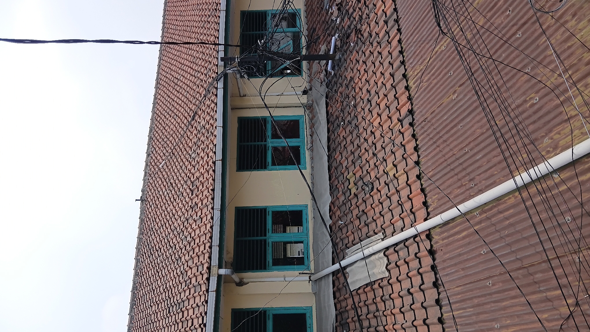 An outdoor, eye-level, medium shot captures a partial view of a building's exterior and rooftops. The building features a red tiled roof and pale yellow walls with three turquoise framed windows, each fitted with horizontal louvers. Below this level, another rooftop is visible, covered with corrugated metal sheets. Numerous black electrical wires crisscross the scene, originating from a black pole positioned on the lower rooftop. A white pipe runs vertically up the building, passing over both rooftops. The sky above is overcast and white. The scene is likely in a densely populated urban area, possibly Bandung City, Indonesia, given the style of the building and the tangled overhead wiring, which is common in many Indonesian cities.