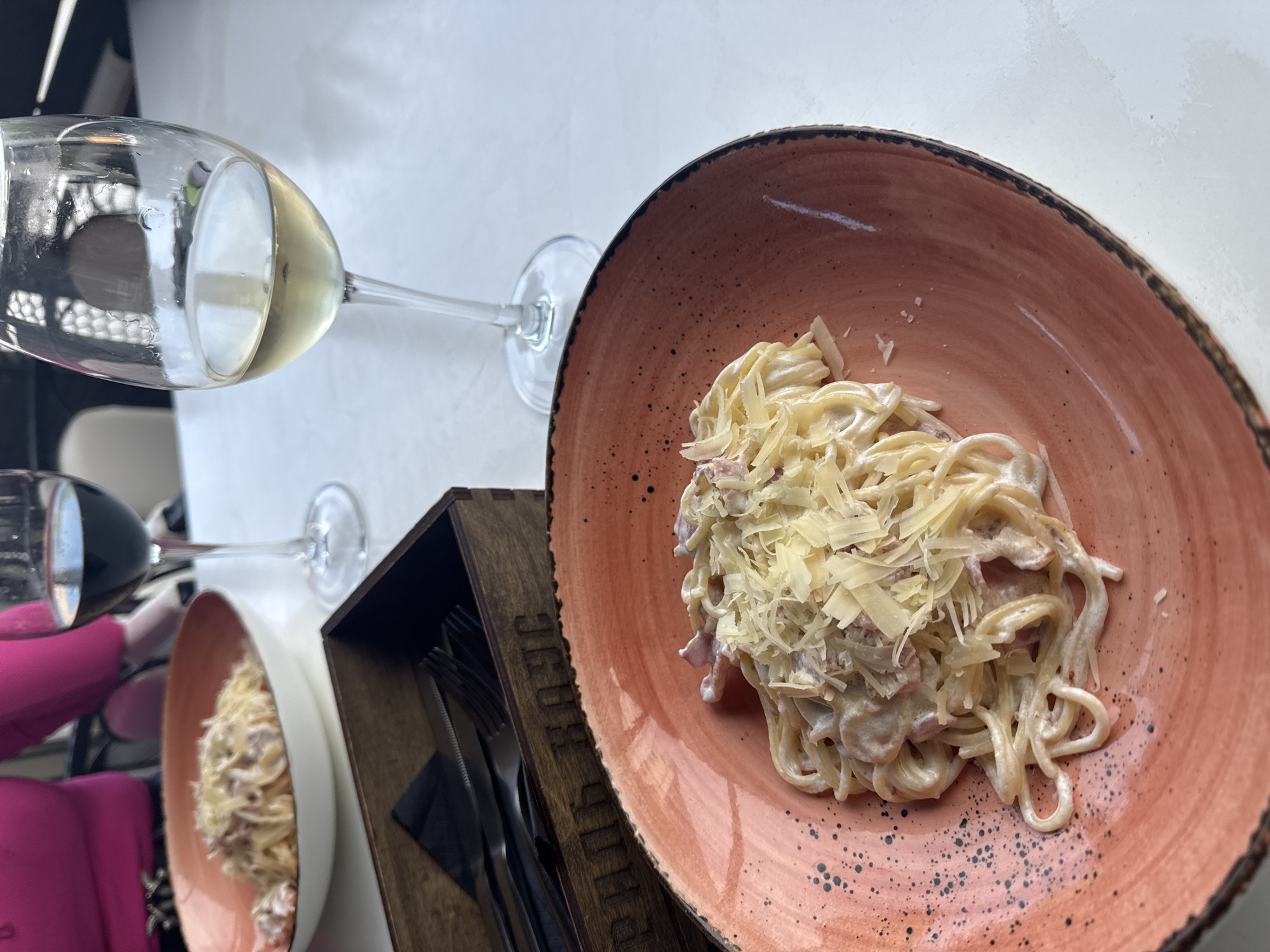 The media file presents an overhead view of a dining scene, likely in a restaurant or cafe in Chaikovsky, Russia, during daytime. The central focus is a generous serving of pasta, appearing to be spaghetti or linguine, served in a distinctive reddish-orange ceramic bowl with a rustic, dark-rimmed edge. The pasta is coated in a creamy white sauce and heavily garnished with shredded or grated hard cheese, with small pieces of meat (possibly bacon or ham) mixed throughout.
On the light-colored table surface, a dark brown wooden box containing silver forks and knives is positioned next to the main pasta dish. Partially visible Cyrillic text is engraved on the side of this wooden box, appearing to spell out "БЕРЛОГА" (Berloga).
Two stemmed wine glasses are present in the frame. One glass, situated in the upper left, holds a pale yellow liquid, indicative of white wine. Further back and to the left, another glass contains a dark red liquid, suggesting red wine. A person's arm, dressed in a bright pink garment, is partially visible near the red wine glass, implying a diner is present. A second, somewhat blurry plate of pasta is also visible in the background, suggesting a meal for two. The scene depicts a casual yet pleasant dining experience.