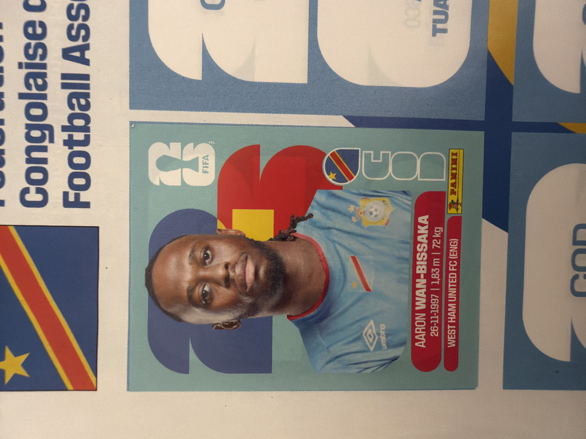 The image is a close-up of a sticker album page. The page features a football player's sticker. The player is Aaron Wan-Bissaka, and the sticker includes his name, date of birth, height, weight, and club, West Ham United FC (ENG). The sticker also displays the Panini logo. The background of the sticker shows a stylized representation of the Democratic Republic of Congo flag and a large number "23". The surrounding area of the album page has larger graphic elements in blue and white, with some text that appears to be in French, reading "Congolaise c Football Ass". There is no information in the image or the provided context that would indicate the location is in the city of Zlatitrap, Bulgaria.