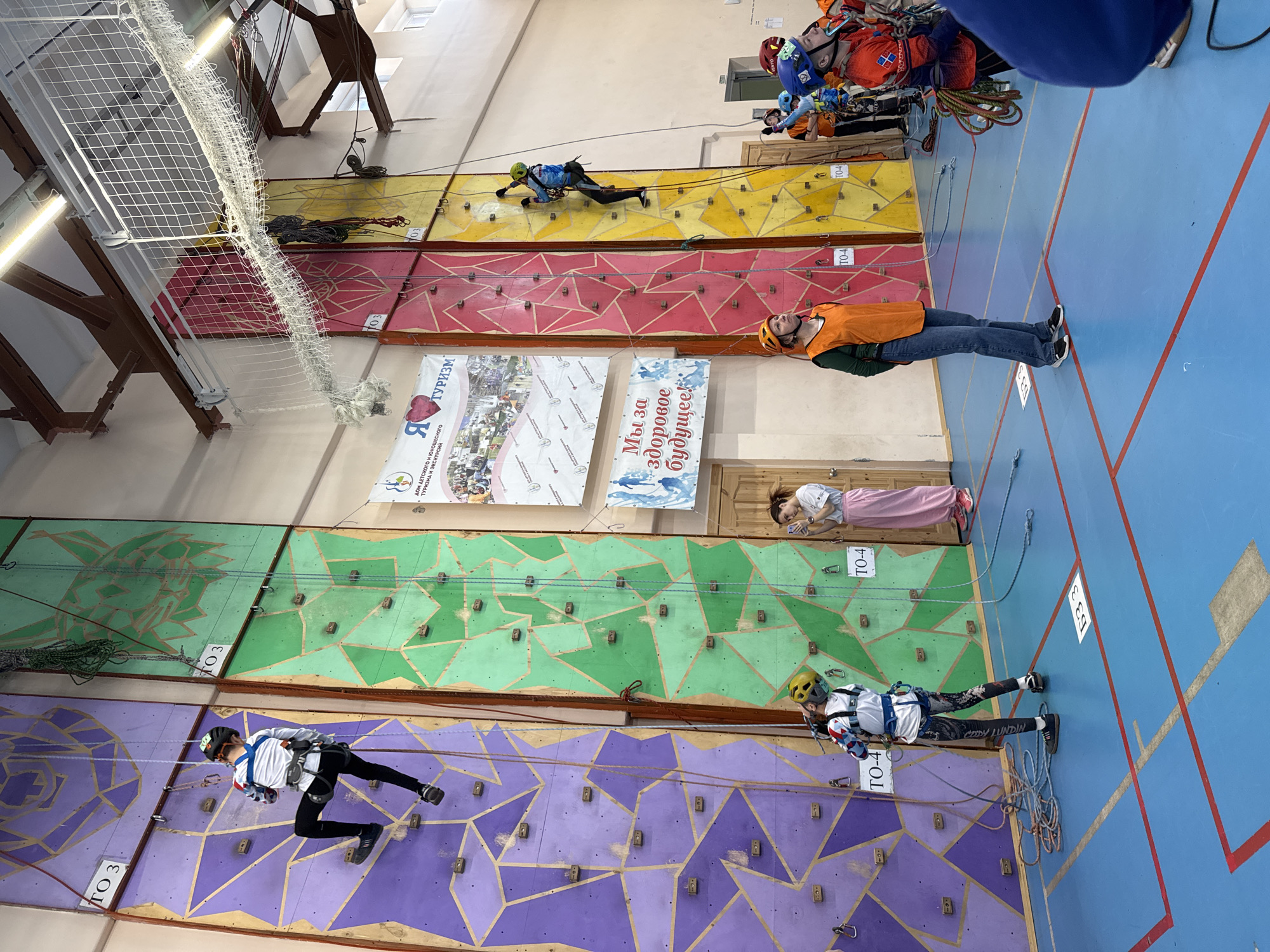 The image shows a climbing gym in Berezniki, Russia. Several people are actively climbing the colorful artificial walls, while others are observing or preparing to climb. The walls are divided into sections marked with numbers. Banners hang on the wall, one advertising tourism and excursions, and another promoting a healthy future. Climbing equipment such as ropes and harnesses are visible. The gym has a blue floor with red and white markings. A net hangs from the ceiling.