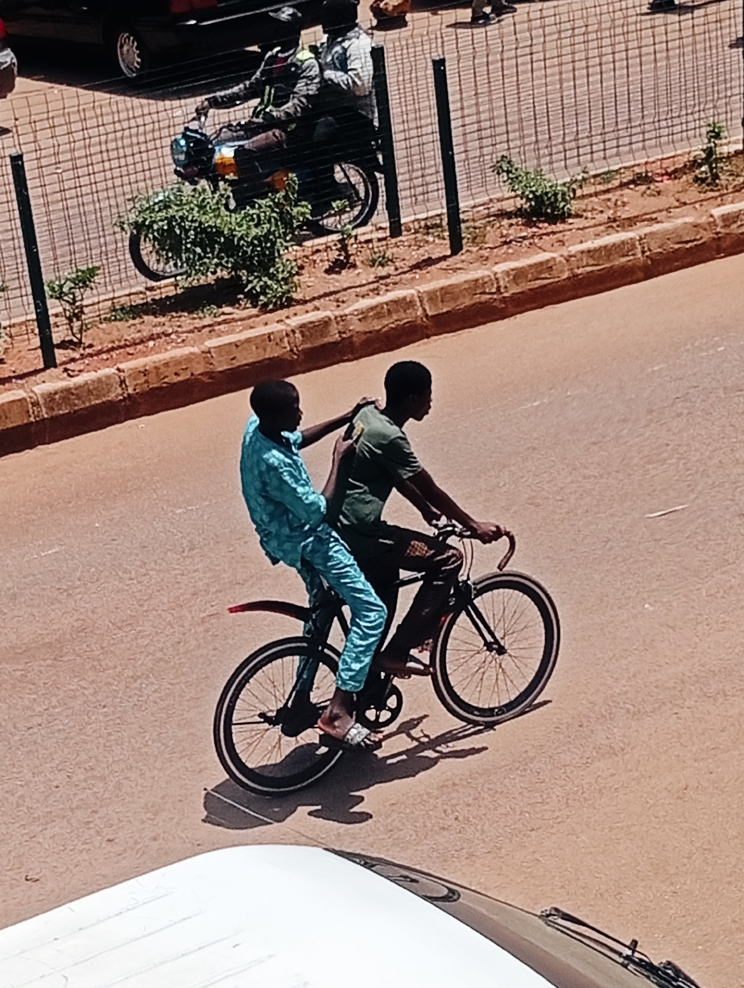 In Bukuru, Nigeria, two individuals are captured riding a bicycle on a paved road. The person in front, wearing a green t-shirt, is pedaling the bicycle, while the person behind, dressed in a blue patterned outfit, is sitting behind them, with their left arm around the rider's shoulder. The bicycle has black tires with silver rims and spokes, and a red mudguard over the rear wheel. The road surface appears to be asphalt, with a lighter-colored line marking the center. To the left of the road, a wire fence with vertical posts separates it from a dirt area with sparse green vegetation. Beyond the fence, a dark-colored vehicle is partially visible, along with a motorcycle carrying two individuals. The scene is illuminated by sunlight, casting shadows on the road.