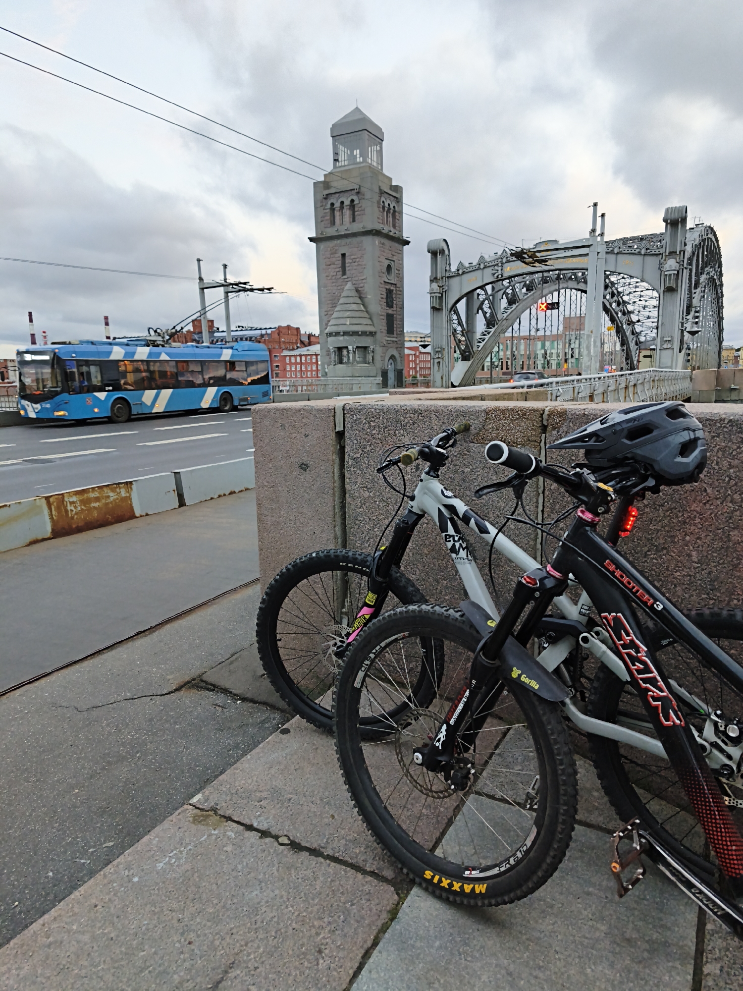 The image shows two mountain bikes parked on a stone bridge in a city. The bikes are leaning against a stone wall. The bike on the right is black with red accents, and has "SHOOTER 3" written on the frame. The bike on the left is grey with black accents. A black helmet is resting on the handlebars of the bike on the right. In the background, a blue and white trolleybus is driving across the bridge. The bridge has a distinctive arched structure. The sky is overcast. The location is Unknown, Unknown.
