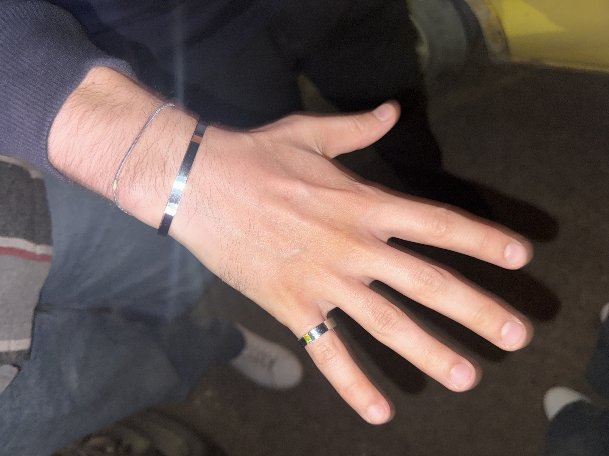 A medium shot depicts a person's left hand adorned with a ring on the pinky finger and two bracelets on the wrist. The hand is fair-skinned with short fingernails. The bracelets are silver and one is thin while the other is thicker. The ring is also silver. The person is wearing a dark-colored long-sleeved shirt and blue jeans. A small section of what appears to be a white sneaker is visible in the background. The location and context of the city of Izhevsk, Russia, are not readily apparent from the image itself.