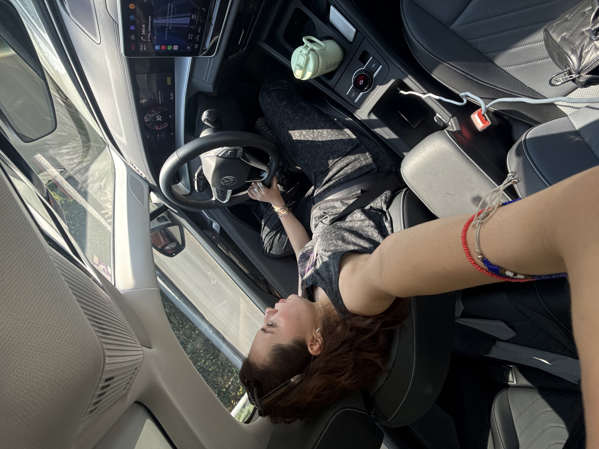 A woman is inside a vehicle, presumably a car, taking a selfie. The vehicle's dashboard includes a digital screen displaying navigation information in Spanish, which mentions "Cale Claveranes," suggesting a location in or near Mexico City. The woman is wearing casual clothes and has her seatbelt on. A water bottle and other items are visible in the car's interior.