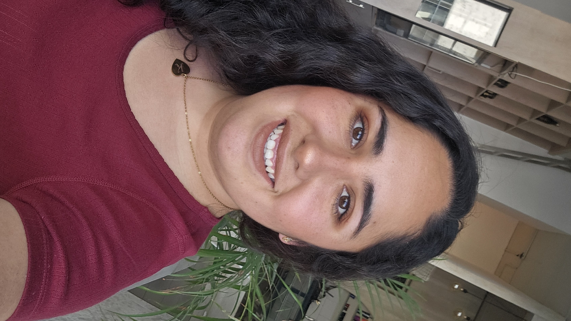 A woman is centered in a portrait shot. She has dark brown hair, brown eyes, and a warm skin tone. She is wearing a maroon shirt and a gold necklace with a heart pendant. The pendant appears to have a letter on it. She is smiling, revealing white teeth. The background is blurred, but a houseplant is visible in the foreground. The location cannot be determined by the visual data provided.