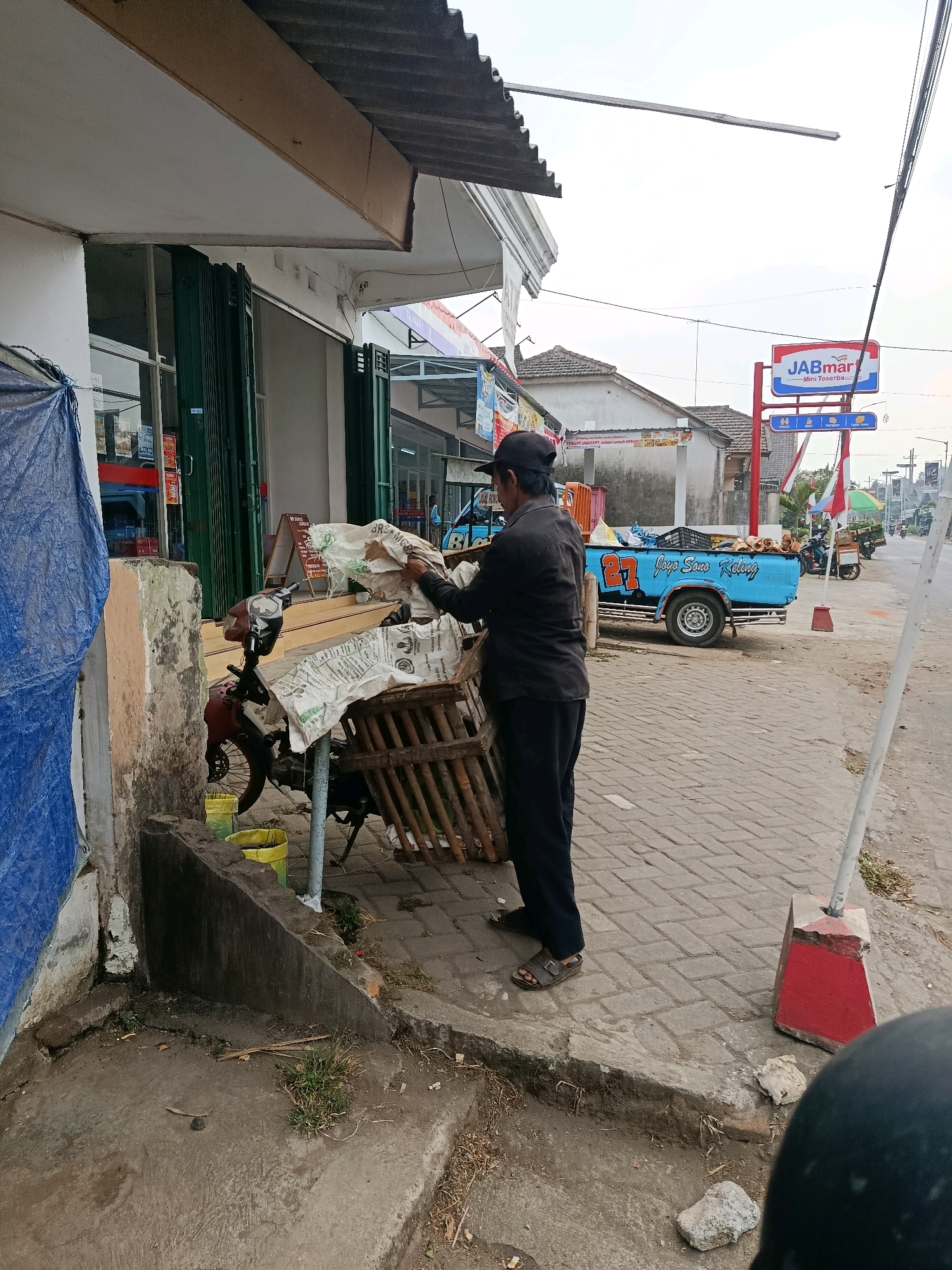 In Tajinan, Indonesia, a man wearing a dark baseball cap, dark long-sleeved shirt, and dark pants stands on a brick-paved sidewalk next to a weathered, light-colored building with a corrugated metal awning. He is handling large white sacks that are partially covering a wooden crate and a red motorcycle. The motorcycle has a black seat and red accents. Yellow buckets are visible near the motorcycle. To the right of the man, a light blue pickup truck with the number "27" and text "Joya Sano Keling" on its side is parked. A red and white sign for "JABmart - Mini Toserba" stands further down the street. In the background, other buildings and parked motorcycles can be seen. The ground is a mixture of paved brick and dirt.