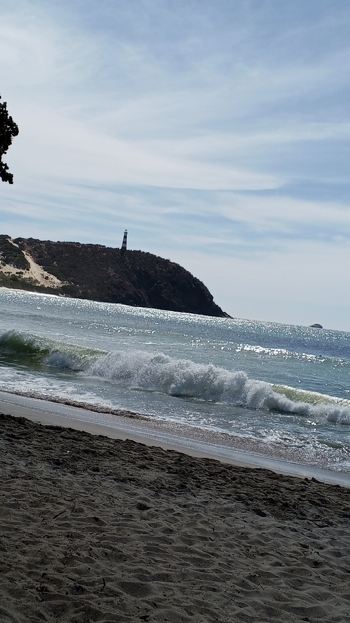 The image depicts a beach scene. In the background, a hill is visible with a lighthouse standing atop it. The sea is in the middle ground, with waves breaking near the shore. The foreground is dominated by dark-colored sand. The sky is partly cloudy. Location is Unknown, Unknown.