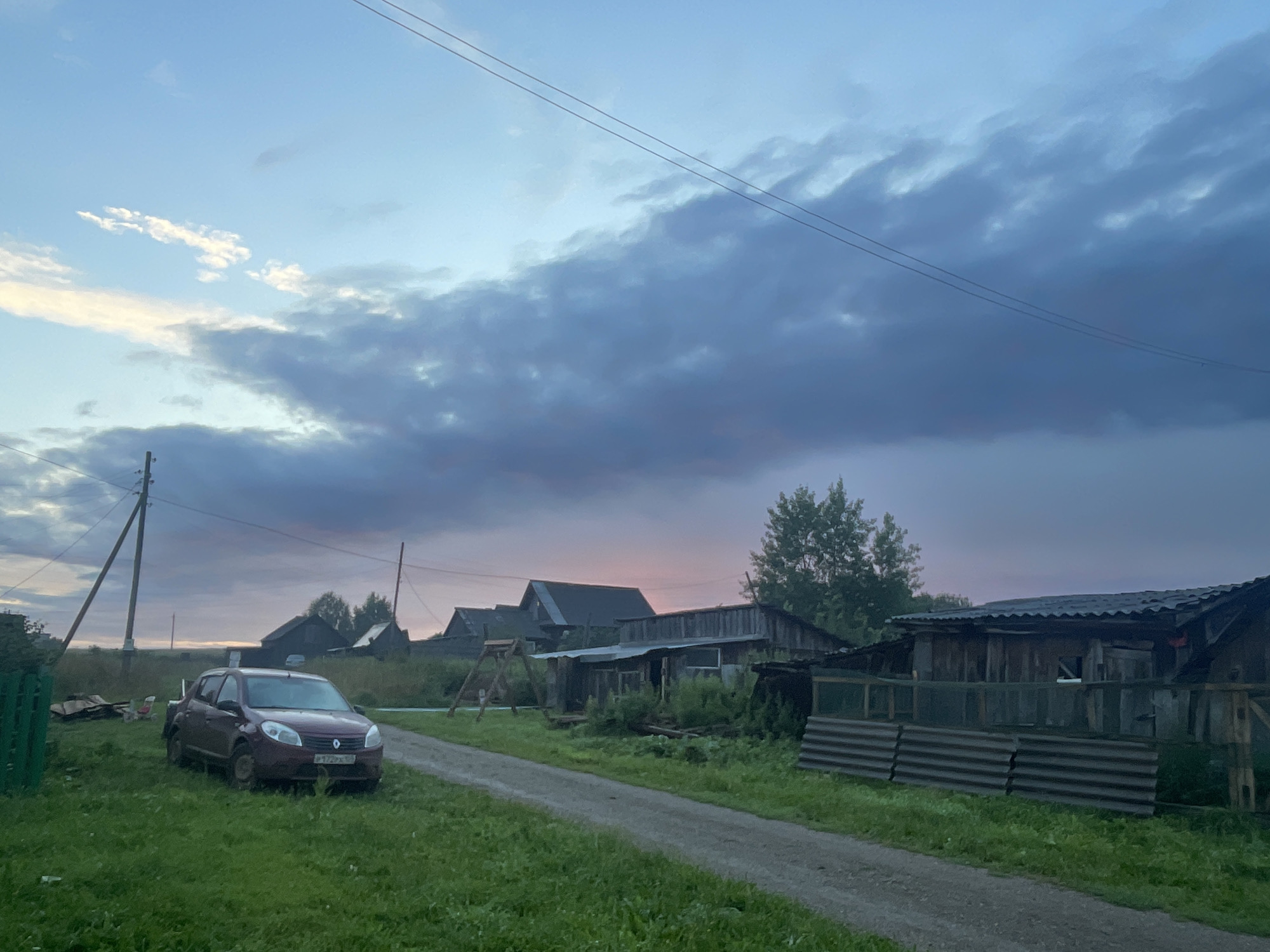 The image captures a rural scene at dusk in Panteleevka, Russia. A dirt road runs through the center of the frame, bordered by green grass and vegetation. On the left side of the road, a maroon sedan is parked on the grass. In the background, several wooden structures, typical of a village, are visible, including houses and sheds. The sky above is filled with dramatic clouds, with hints of pink and orange suggesting the setting sun. Power lines are strung across the upper portion of the image, supported by wooden poles. The overall atmosphere is peaceful and serene, indicative of a quiet evening in the countryside.