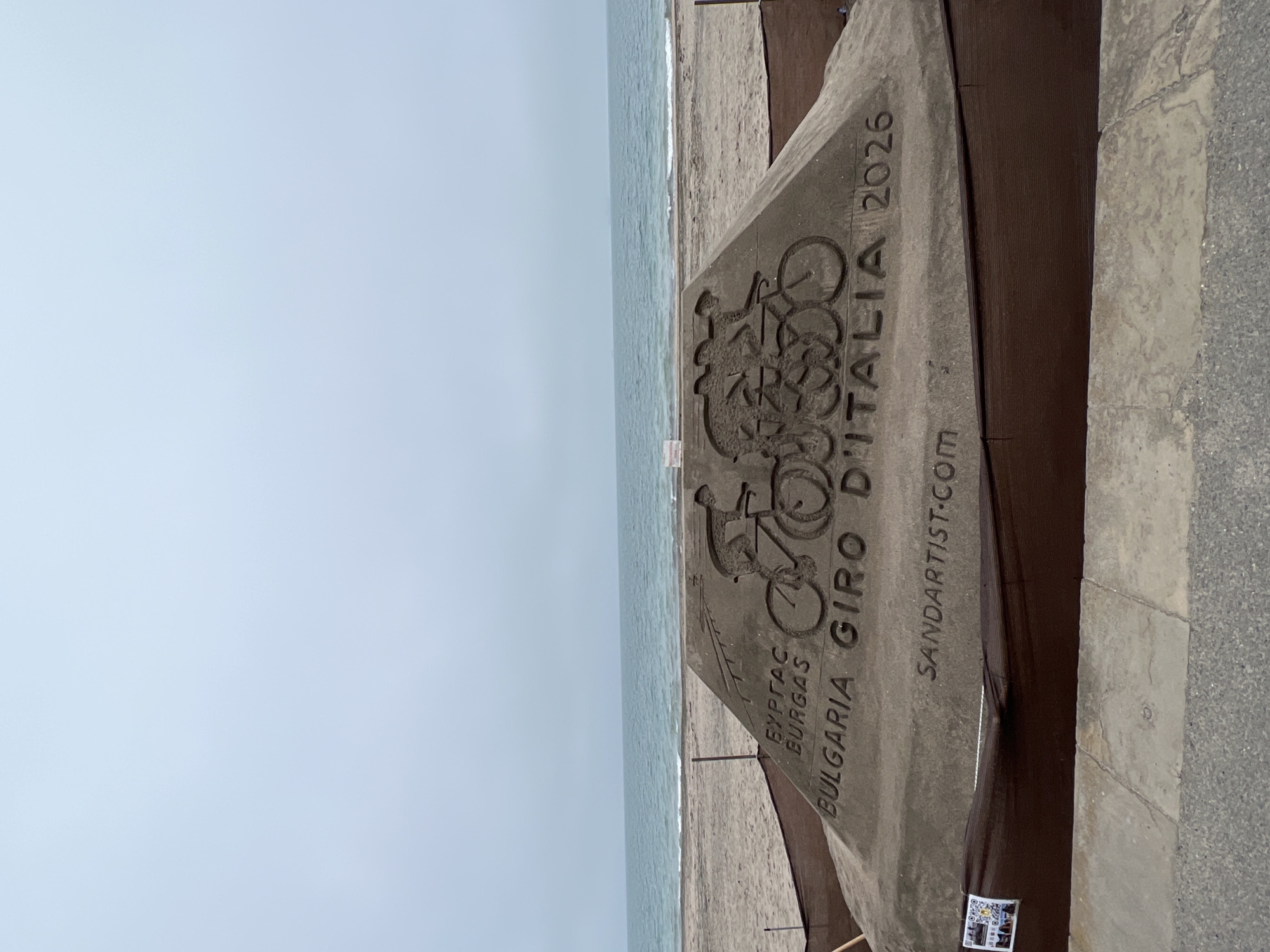 A sideways view shows a sand sculpture on a beach in Burgas, Bulgaria. The sculpture is dedicated to the Giro D'Italia 2026 cycling event. It includes images of bicycles and the text "BULGARIA BURGAS GIRO D'ITALIA 2026 SANDARTIST.com." The Black Sea is visible in the background under a cloudy sky. The sculpture is situated next to a stone walkway or retaining wall. A QR code is affixed to the base of the sculpture.
