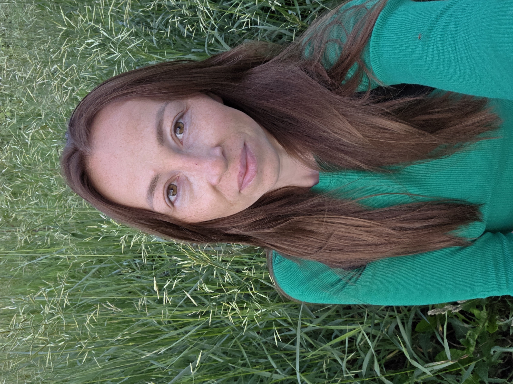 A woman with long brown hair and freckles is taking a selfie outdoors in front of a field of tall green grass. She is wearing a bright green ribbed long-sleeved shirt. Her eyes are light brown, and she is looking directly at the camera with a neutral expression. The background is a dense field of grass, with some seed heads visible. The lighting suggests it is daytime, possibly late afternoon or early evening, given the soft light. There is no indication of the city of Chaikovsky, Russia, or any specific urban elements in the image.