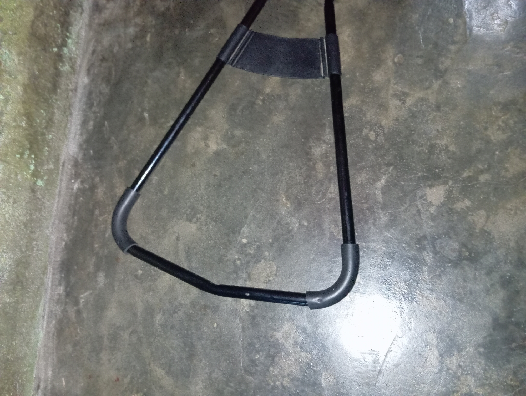 A black metal exercise device, likely an ab roller or similar core strengthening equipment, is positioned on a weathered concrete surface. The device has a triangular frame made of black tubing, with grey rubberized grips on the two lower corners and one upper corner. A black plastic component is attached to the upper right side of the frame, serving as a stabilizer or handle. The concrete floor appears to be outdoors, possibly in a courtyard or alleyway in Rejosari, Indonesia, given the texture and color of the surface and the visible mossy growth on a wall in the background. The lighting suggests it is daytime, with a strong light source reflecting off the wet or polished concrete in the lower left portion of the image. There are no people or other living beings visible in the scene.