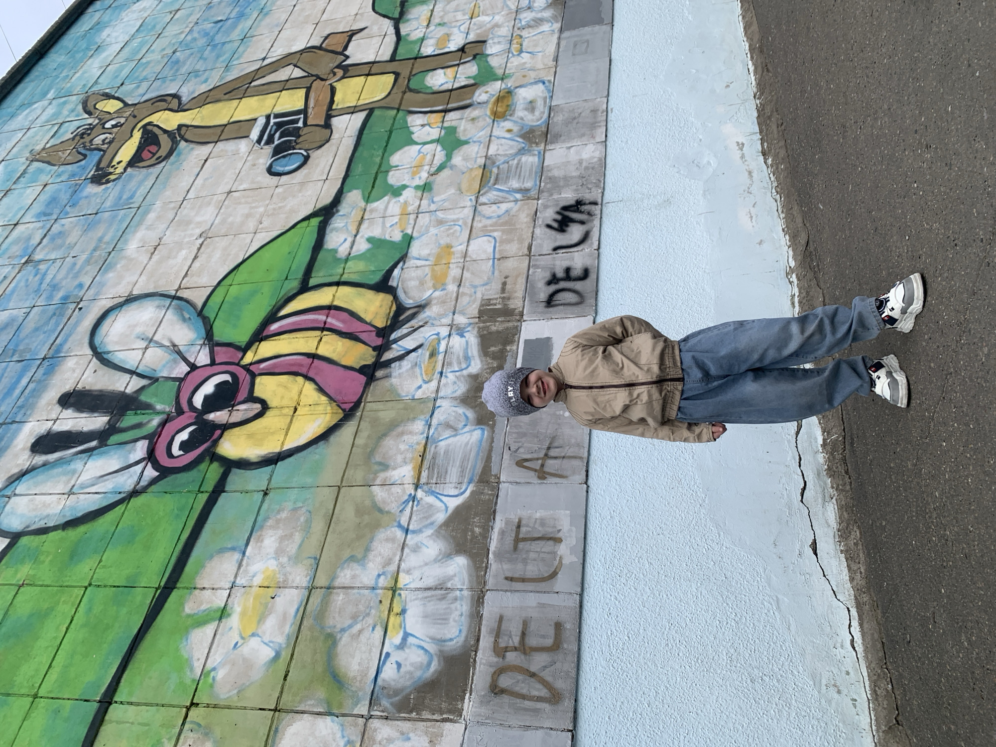 A young boy stands beside a tiled mural in Severodvinsk, Russia. The mural features a cartoon squirrel with a camera, a bee, and flowers. The word "DELTA" is written in graffiti on the tiles below the mural. The boy wears a beige jacket, blue jeans, a grey hat, and white sneakers. A cracked sidewalk runs parallel to the mural.