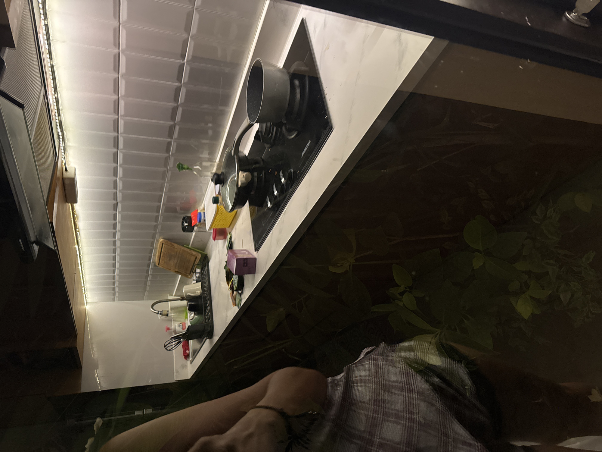 The image depicts a kitchen scene seemingly reflected in a dark window or glass surface, with the reflection superimposed over the foreground.
The kitchen features white subway tile backsplash, a black stovetop with a silver pot, and a countertop. A sink with a drying rack and various kitchen utensils are visible.
In the foreground, a person is discernible, wearing a checkered shirt. Part of their arm is visible, suggesting they may be near the window.
The reflected environment appears to be a dark outdoor area filled with foliage.
The prompt mentions Jimbaran, Indonesia, but nothing in the visual data explicitly identifies the location.