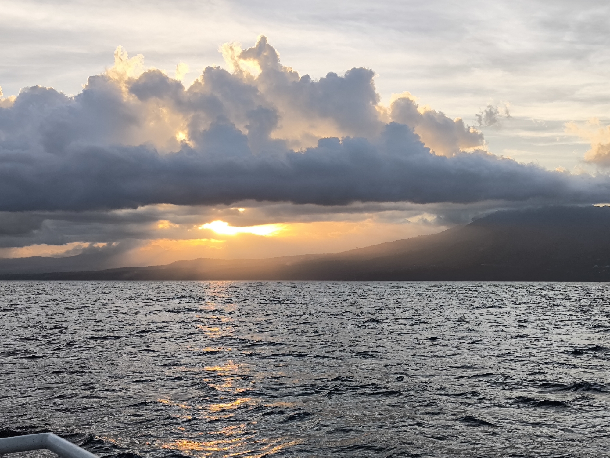 The image depicts a body of water, likely a sea or large lake, under a sky with dramatic clouds at sunset or sunrise. The sun is obscured by a thick layer of dark clouds, but its rays are visible, breaking through the cloud cover and illuminating the water and a distant landmass. The landmass appears to be a mountainous area with a coastline. The water is rippled, reflecting the golden light of the sun. The foreground of the image is slightly obscured, with a glimpse of what appears to be a railing or part of a boat, indicating the photo was taken from a vessel. The location is Unknown, Indonesia.