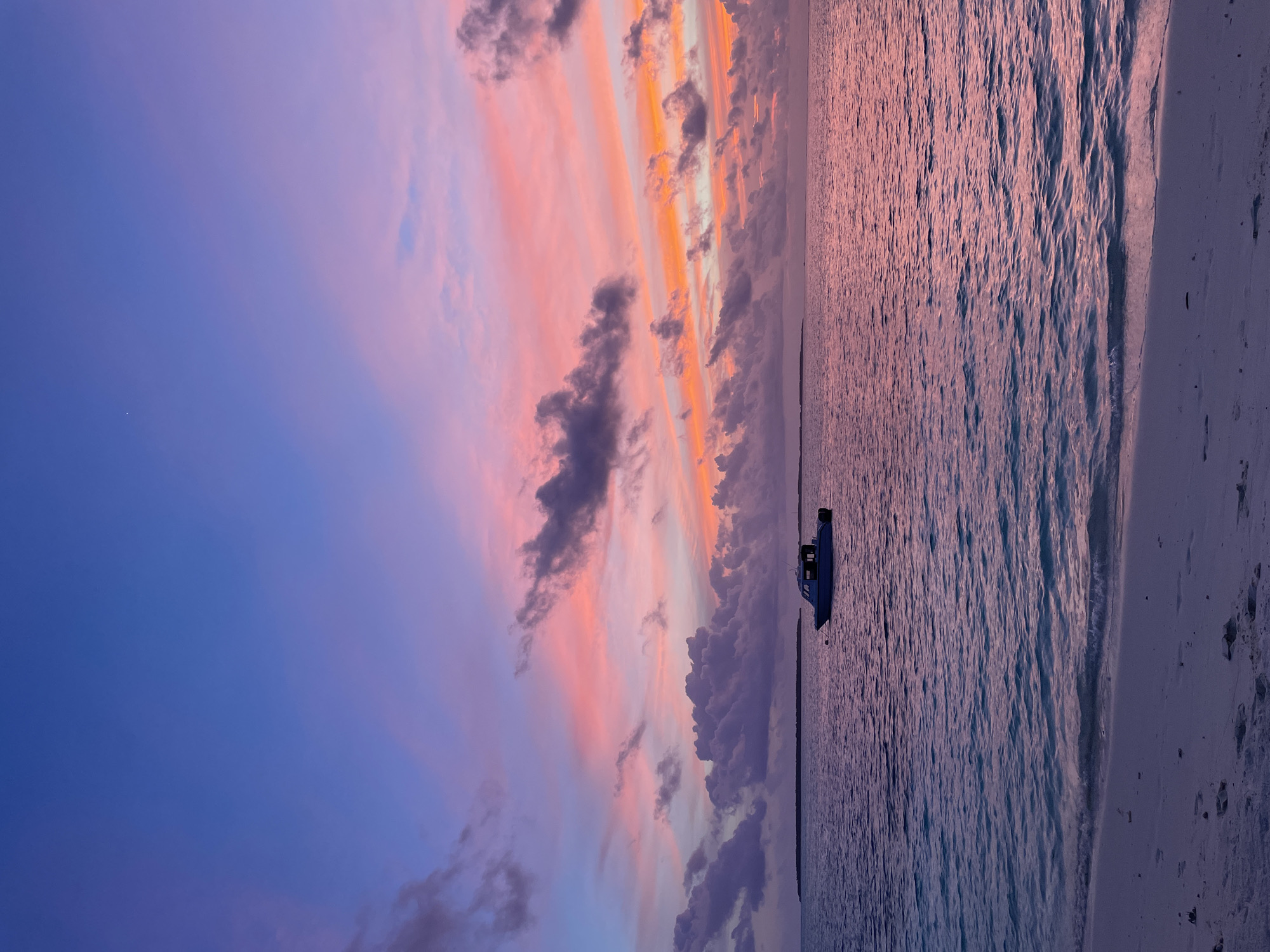 The image depicts a serene seascape at dusk. The sky is a vibrant canvas of color, transitioning from a deep blue at the top to shades of pink, orange, and purple towards the horizon. Scattered clouds add texture and depth to the sky. In the foreground, a calm body of water reflects the colors of the sunset. A small boat is visible in the middle ground, anchored on the water. The shoreline in the immediate foreground consists of light-colored sand with some footprints visible. In the distance, a low-lying island or landmass can be seen. The overall mood of the image is tranquil and picturesque.