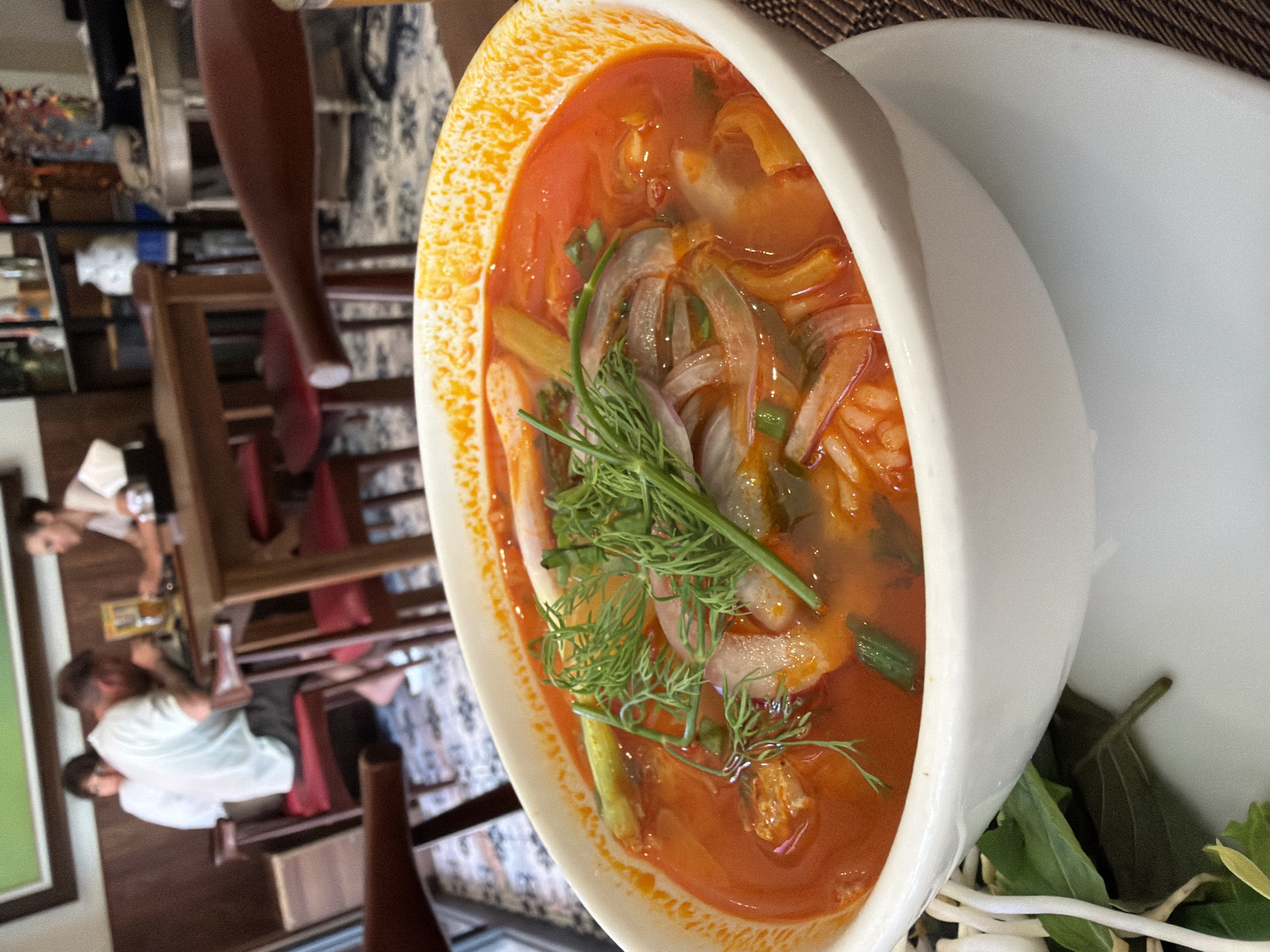 A close-up, eye-level shot shows a steaming bowl of Vietnamese noodle soup, likely Pho, in a white ceramic bowl. The broth is a vibrant red-orange, with visible chunks of meat, sliced onions, and green herbs, including dill and what appear to be scallions. The soup is served with a side of fresh bean sprouts and leafy greens. In the blurred background, two people are seated at a wooden table, with a third person further back. They appear to be in a restaurant setting, indicated by the wooden furniture, patterned floor, and shelves filled with decorative items. The restaurant's ambiance suggests it is located in a place with a tropical climate, potentially Nha Trang Ward, Vietnam, given the style of the food and decor.