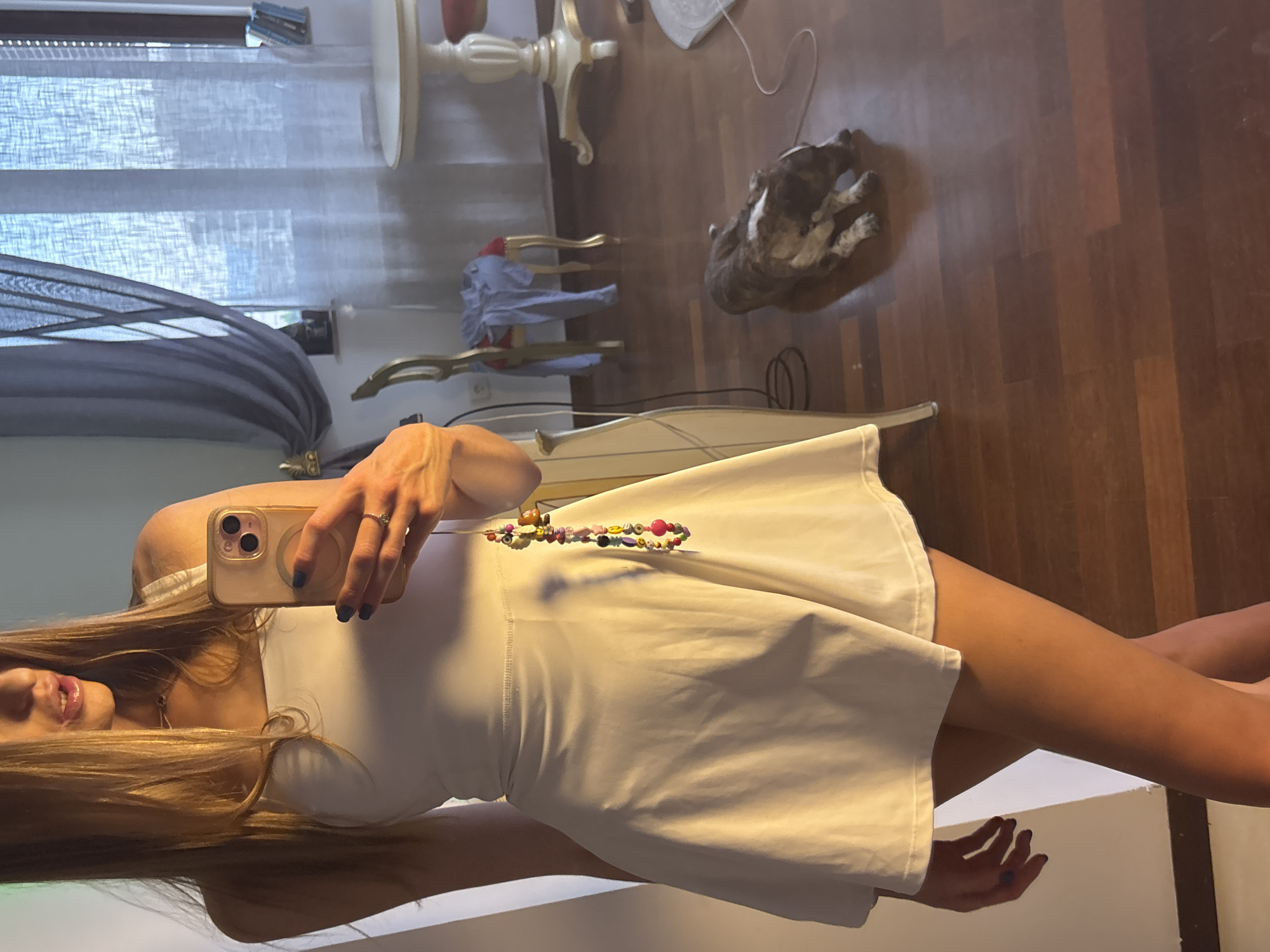 A woman, seen from the waist down and reflected in a mirror, stands in a room. She is wearing a white tank top and a white flared skirt. Her right hand, holding a smartphone, is visible, and she is wearing a ring. A beaded bracelet hangs from her hand. A dog lies on the wooden floor in the background, near a draped chair and a small table. The room has curtains and a window.