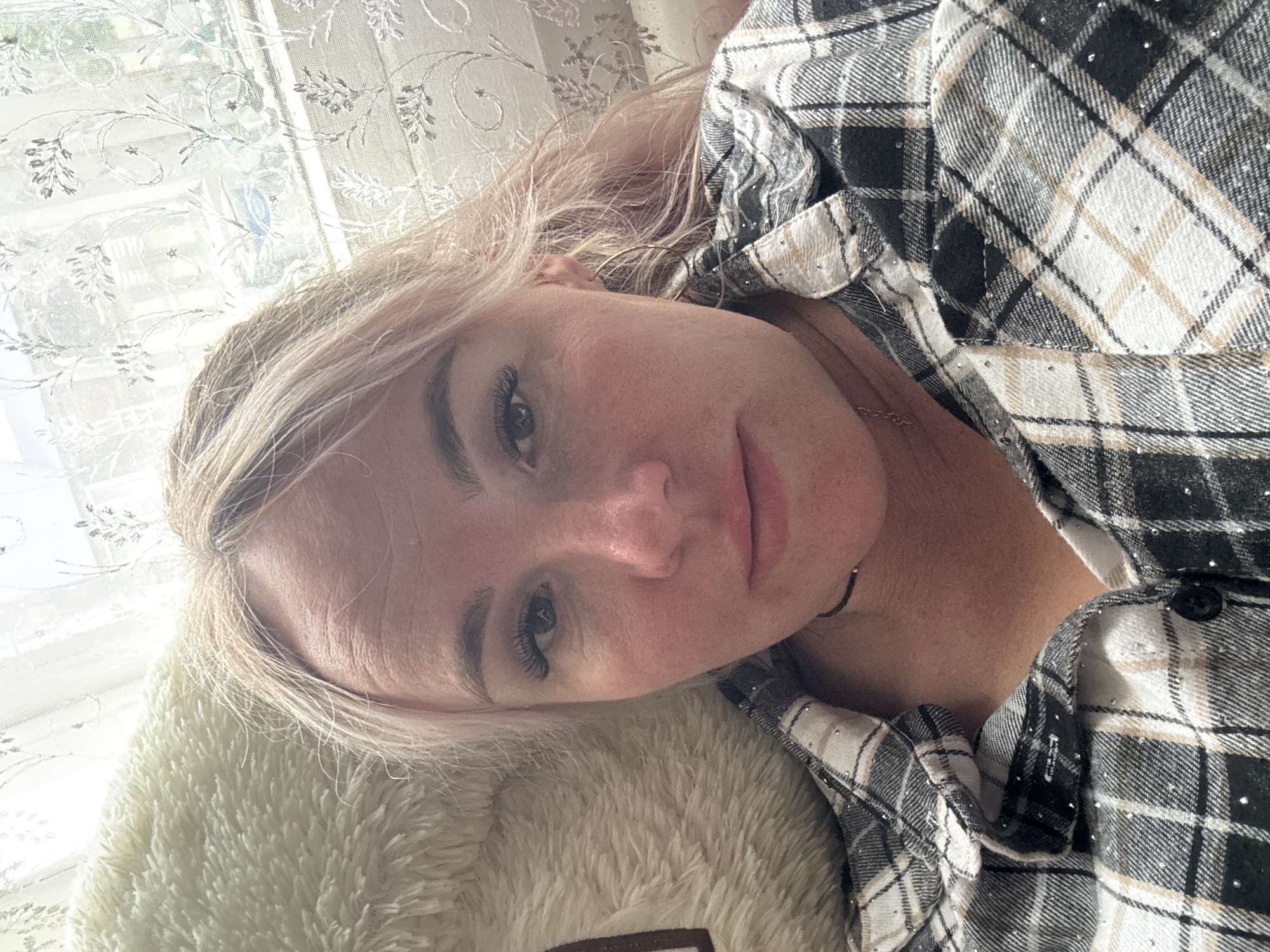 This is a close-up, portrait-style photograph of a woman lying down, with her head resting on a fluffy, cream-colored object. She is wearing a black, white, and tan plaid shirt with subtle sparkling embellishments. The shirt is open at the collar, revealing her neck and a thin necklace. The woman has blonde hair, which is slightly disheveled and framing her face. Her eyes are open, and she appears to be looking upwards or slightly to the side. Her makeup includes prominent eyelashes and defined eyebrows.
The background is out of focus but appears to be a window with a sheer, patterned curtain, suggesting an indoor setting. The lighting suggests it could be daytime, possibly morning or afternoon, with natural light filtering through the window. The overall mood is relaxed and casual. There is no visible text in the image. The location context provided is Pozdyshki, Russia.