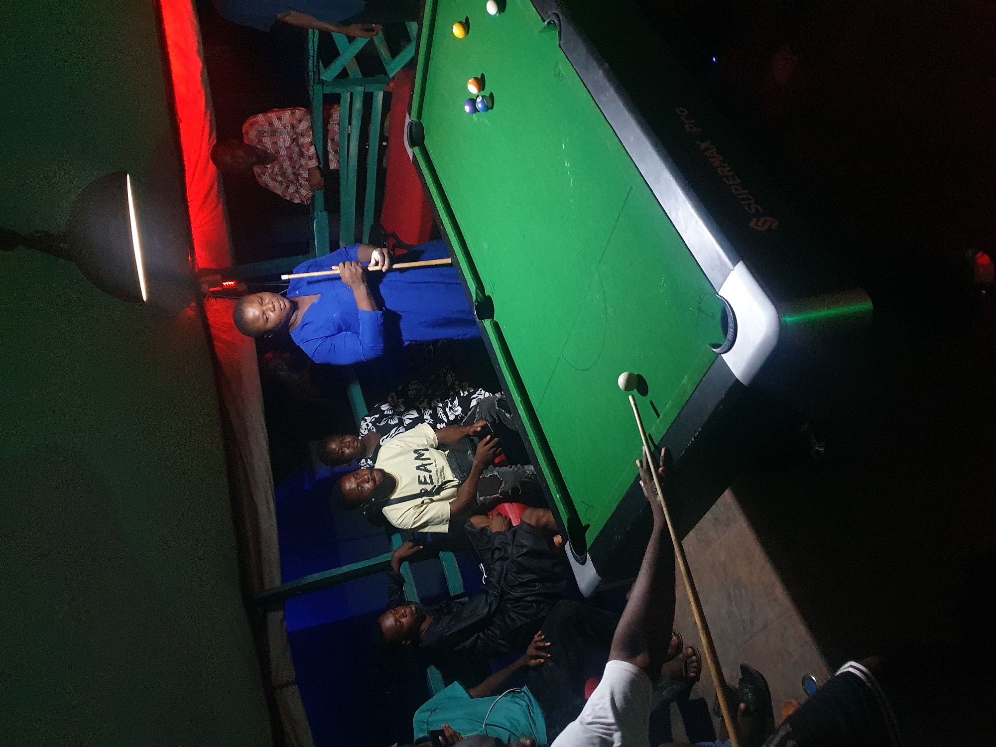 In a dimly lit establishment in Lokoja, Nigeria, a group of people are gathered around a pool table. One woman in a blue dress is preparing to take a shot, while another man is poised to strike the cue ball. The pool table, a "SUPERMAX PRO" model, features a green playing surface with several balls clustered near the center. Several onlookers are seated on benches and chairs surrounding the table, casually observing the game. The room is illuminated by overhead lighting, and the atmosphere appears relaxed.