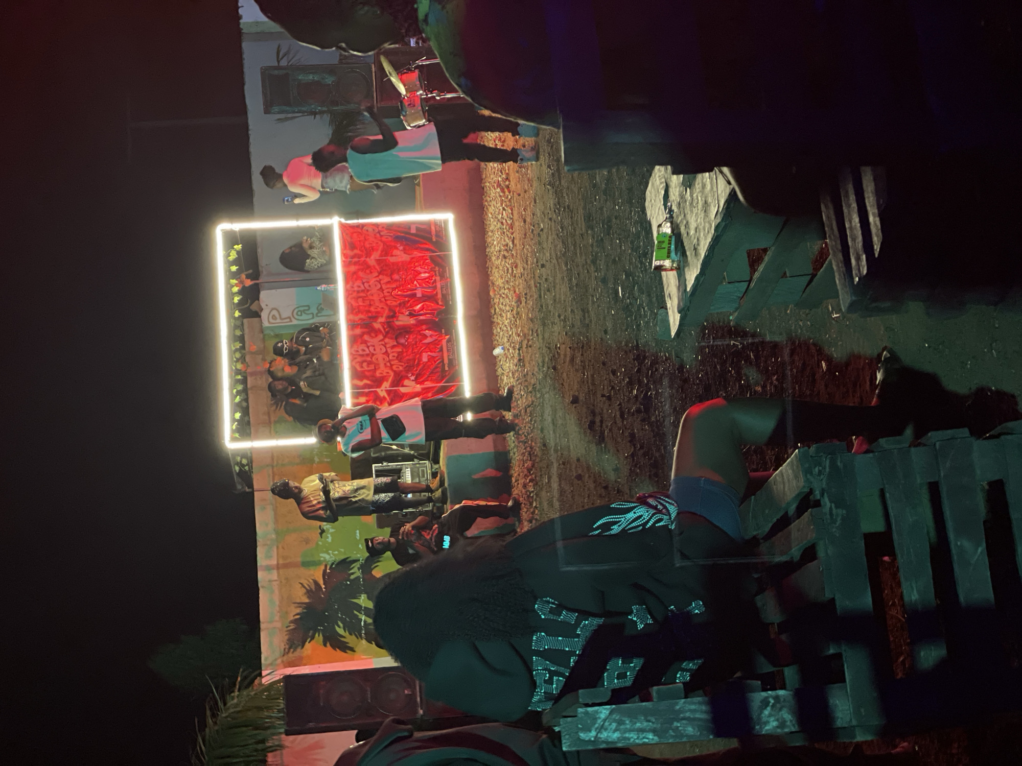 The image captures an outdoor event at night, possibly a block party or concert, in Durumi, Nigeria. The scene is illuminated by artificial lighting, including a prominent neon-lit frame surrounding a stage area. Several people are visible on and around the stage, with some appearing to be performers. One individual is behind a drum set, suggesting live music is taking place. A DJ setup is also present. In the foreground, a person is seated on a makeshift wooden bench, facing away from the camera, observing the event. Another wooden structure, likely a table, is visible to the right with a green bottle and some debris on it. The ground is unpaved and appears to be made of gravel and dirt. In the background, a wall with a mural depicting palm trees and colorful patterns is visible. The overall atmosphere suggests a lively and informal gathering.