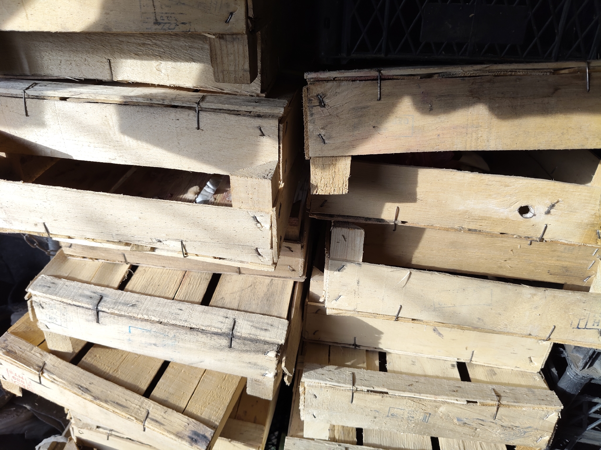 The image shows a stack of weathered wooden crates, some appearing to be damaged. The crates are made of unfinished wood planks, held together with nails. The texture of the wood is rough and shows signs of age and exposure. Sunlight casts shadows across the crates, highlighting their uneven surfaces. The scene is likely an outdoor storage area or a marketplace. There are no people visible in the image, and no specific actions or interactions are depicted. The setting appears to be industrial or agricultural, given the utilitarian nature of the crates. The location is identified as Gîrbova, Moldova, suggesting a rural or semi-urban environment.