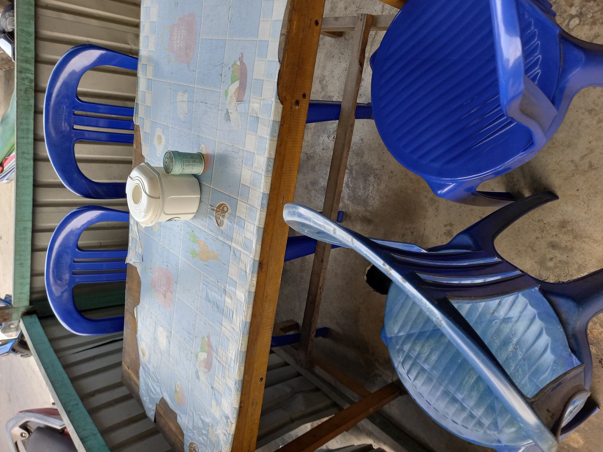 This is a medium shot, eye-level photograph of a small, covered outdoor seating area, likely in Samarinda, Indonesia. The scene depicts a simple wooden table with a blue and white checkered tablecloth, adorned with cartoonish images of fruits and vegetables. On the table, there are two objects: a white cylindrical container with a lid, possibly for tissues or condiments, and a small green cylindrical container, perhaps for toothpicks or sugar.
Four blue plastic chairs are arranged around the table. Two chairs are positioned behind the table, and two are in front, with one of the front chairs angled towards the viewer. The seating area is enclosed by a corrugated metal wall on the left and back, and a light green wooden frame along the top. In the background, the front of a motorcycle, partially visible, suggests an urban or semi-urban setting. The ground is a concrete surface. The lighting is natural, indicating it is daytime, and the overall impression is of a modest, utilitarian space for casual dining or resting.