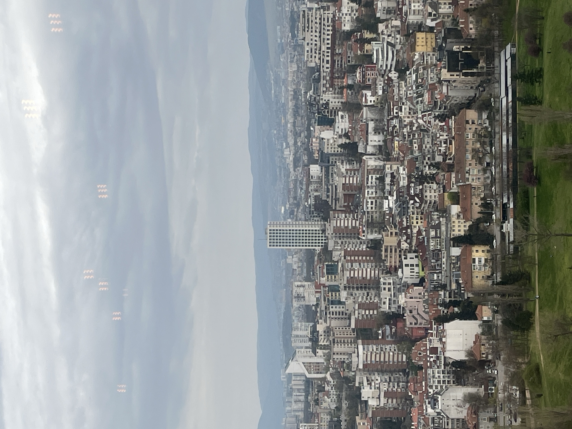 The image shows an aerial view of a densely populated urban area, with a mix of residential and commercial buildings, likely taken in Sofia, Bulgaria. The buildings are primarily low to mid-rise, varying in color and architectural style. A prominent, taller rectangular building with a grid-like facade is visible. In the background, mountains are visible under a cloudy sky. A green park area runs alongside some buildings on the right. On the left, patterns of light are visible in the sky. There is no evidence of direct human activity.