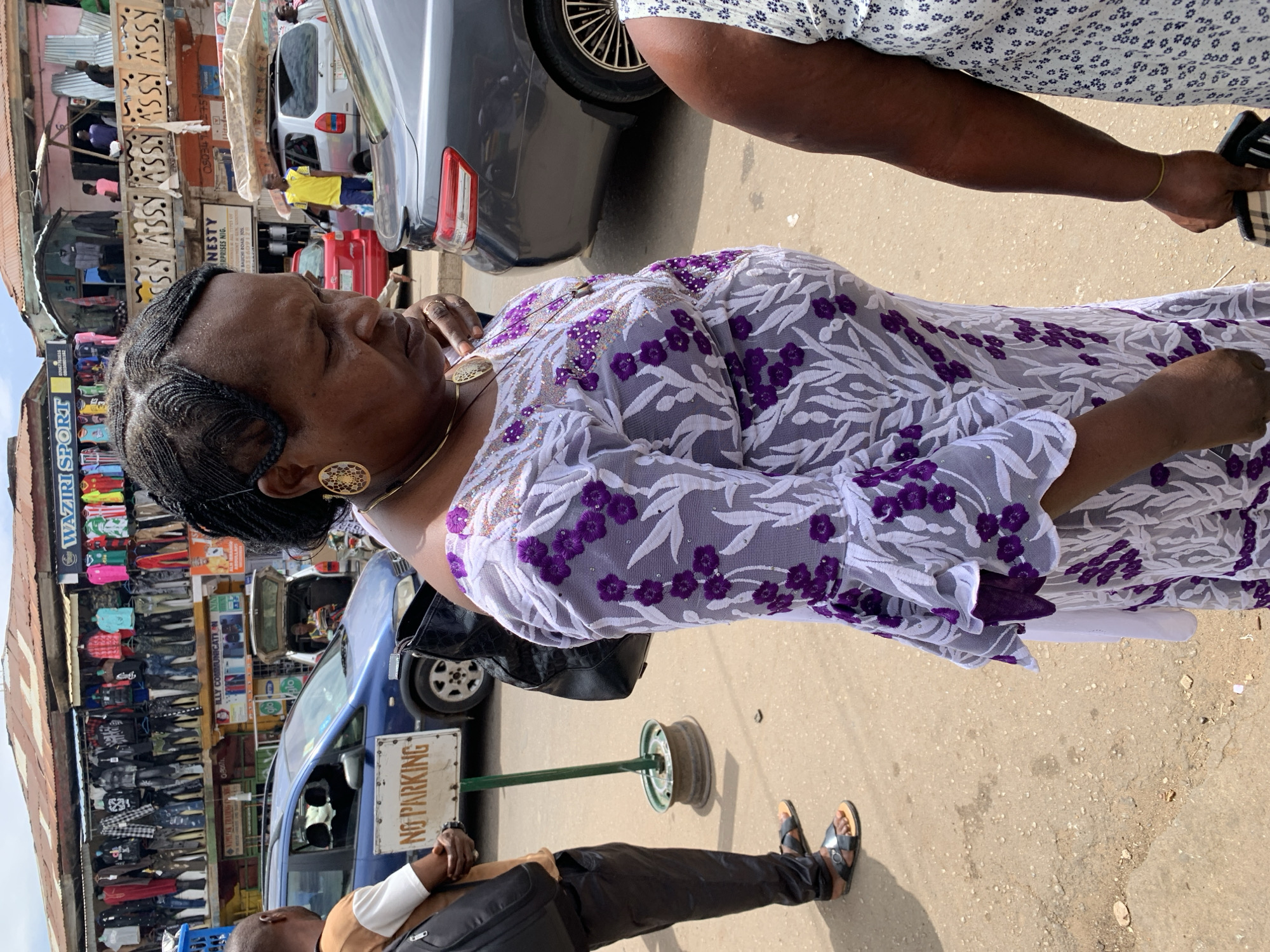 The image depicts an African woman in Jos, Nigeria. She is wearing a lavender and white lace dress with purple floral patterns, gold earrings, and a gold necklace. Her hair is styled in braids. She is standing on a paved street with a "NO PARKING" sign nearby. In the background, there is a building with a sign that reads "WAZIRI SPORT" and "ILLY COMMUNICATI". Several cars and people are also visible in the background. A man stands in front of the "NO PARKING" sign, and another person is partially visible to the right of the woman.