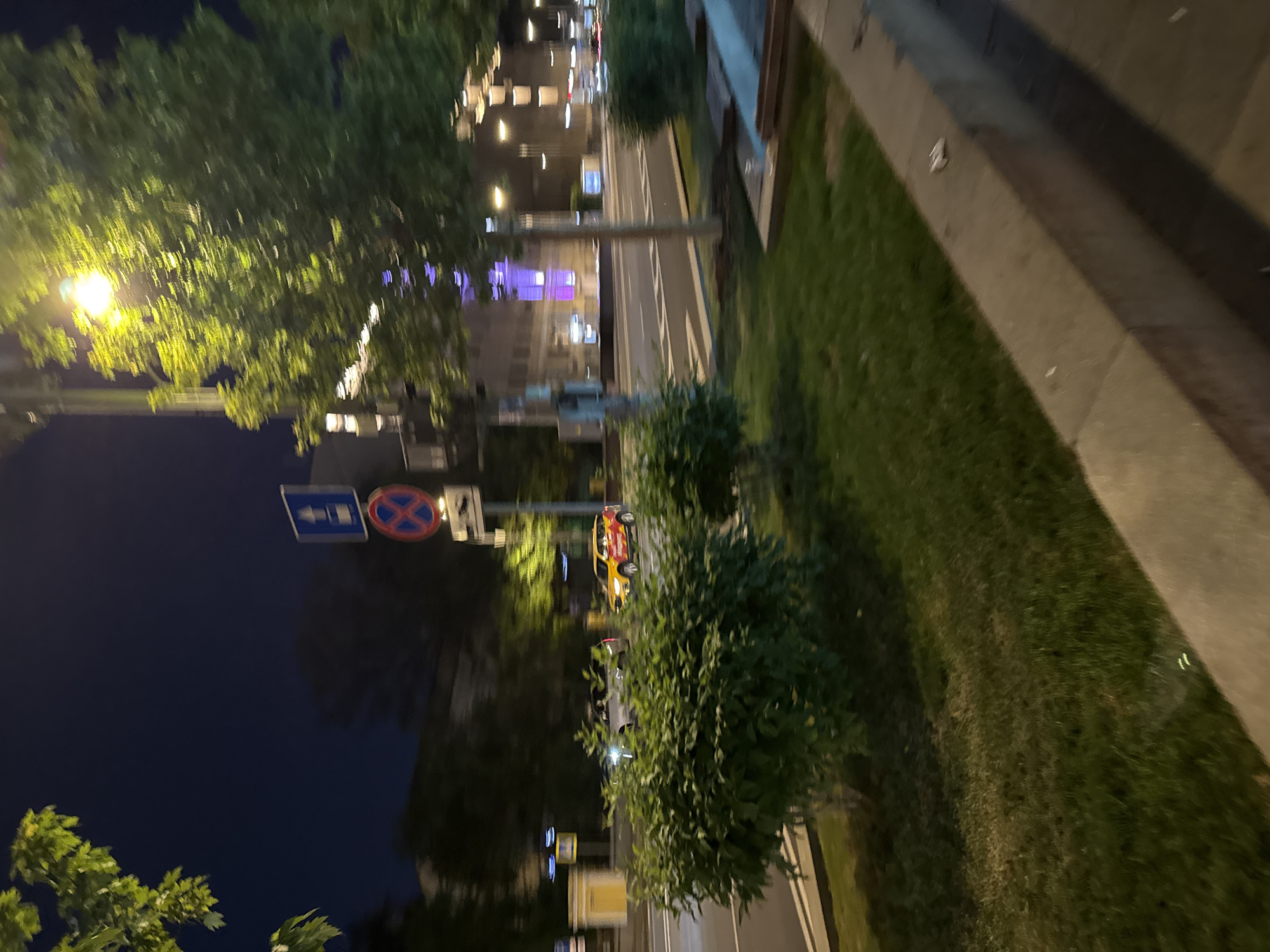 This image captures a nighttime urban scene along Tverskaya Street in Moscow, Russia, with the distinctively illuminated Central Telegraph building visible in the background. The perspective is from a raised sidewalk or median, looking along the multi-lane thoroughfare.
In the immediate foreground on the right, a concrete curb or low wall delineates the edge of a paved area from a grassy strip. This strip features several well-maintained green bushes. In the midground, the asphalt road consists of multiple lanes, marked with white painted lines, and appears to carry traffic in one direction. A central median strip, also covered in green bushes and trees, separates the road.
Two vehicles are visible in motion on the street, exhibiting slight motion blur. The closest is a light-colored SUV with illuminated headlights, traveling from left to right. Further down the street, a yellow and red branded car, identifiable as a Yandex.Taxi, also with illuminated headlights, moves in the same direction.
Road signage is present on a pole near the center-right of the image. A blue rectangular sign with a white upward arrow indicates a required direction or one-way street, positioned above a red circular sign with a blue diagonal cross, signifying "No Stopping" or "No Waiting."
The scene is illuminated by numerous artificial light sources. Bright streetlights with a warm hue cast light on the trees and road. In the background, on the far side of Tverskaya Street, several multi-story buildings are visible. One prominent building in the mid-distance is distinctly illuminated with purple or blue accent lighting, strongly identifying it as the Central Telegraph building. The sky above is dark, indicative of night.