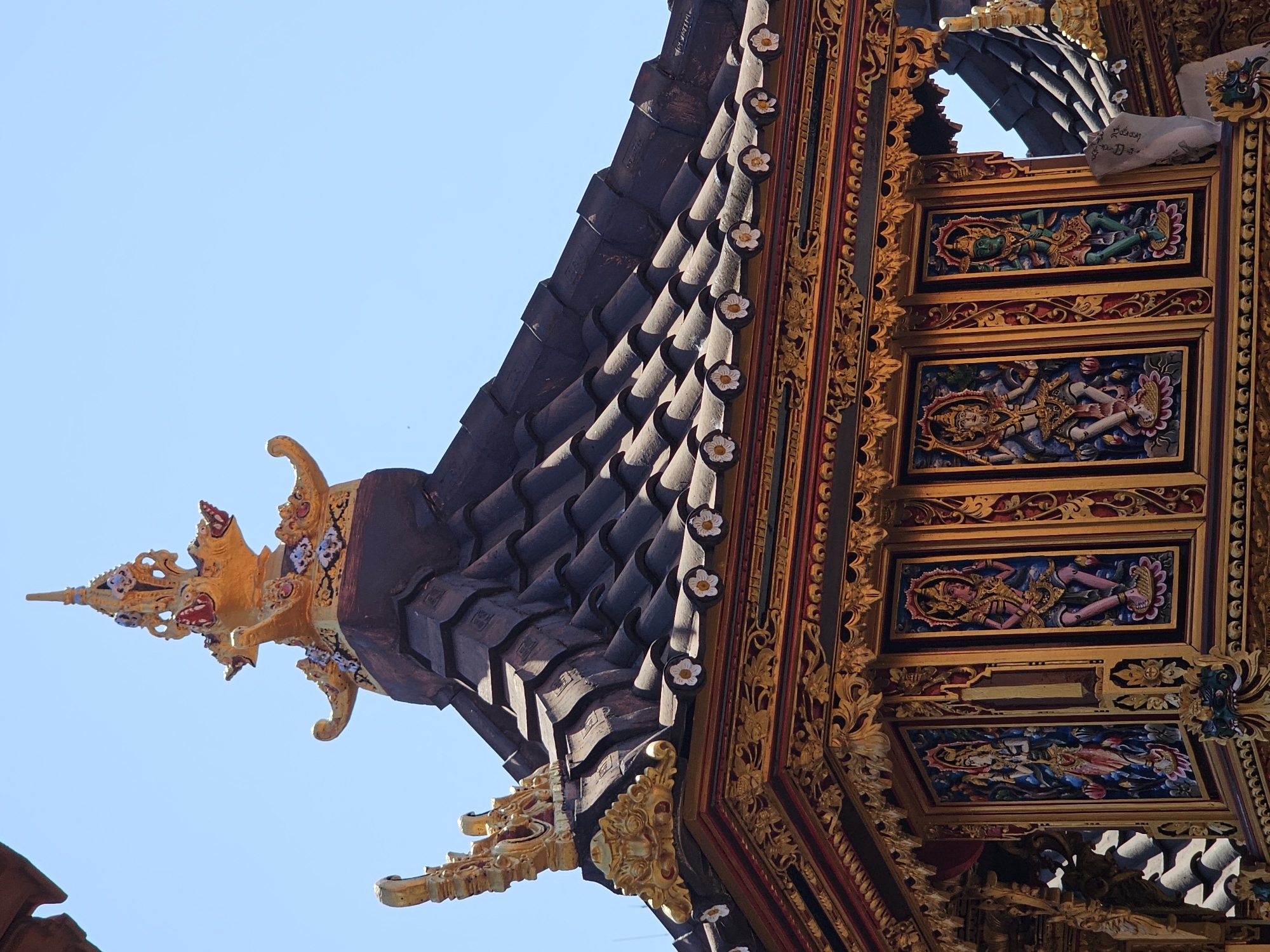 The image captures an ornate, tiered temple roof against a clear blue sky. The roof is covered in dark, overlapping tiles. Along the eaves, intricate golden decorative elements are prominent, including detailed carvings and small white floral motifs. Below the eaves, the sides of the structure are adorned with framed paintings of figures, predominantly in vibrant colors. These figures appear to be deities or mythical beings, depicted in elaborate costumes and postures. The golden trim is extensive, framing these painted panels and accentuating the architectural details. The top of the roof features a multi-tiered golden spire with ornate, stylized dragon-like heads and intricate patterns. This architectural style is characteristic of Balinese temples, suggesting the location is likely within Bali, Indonesia. Given the details of the temple architecture, it is plausible this is a structure within the city of Denpasar.