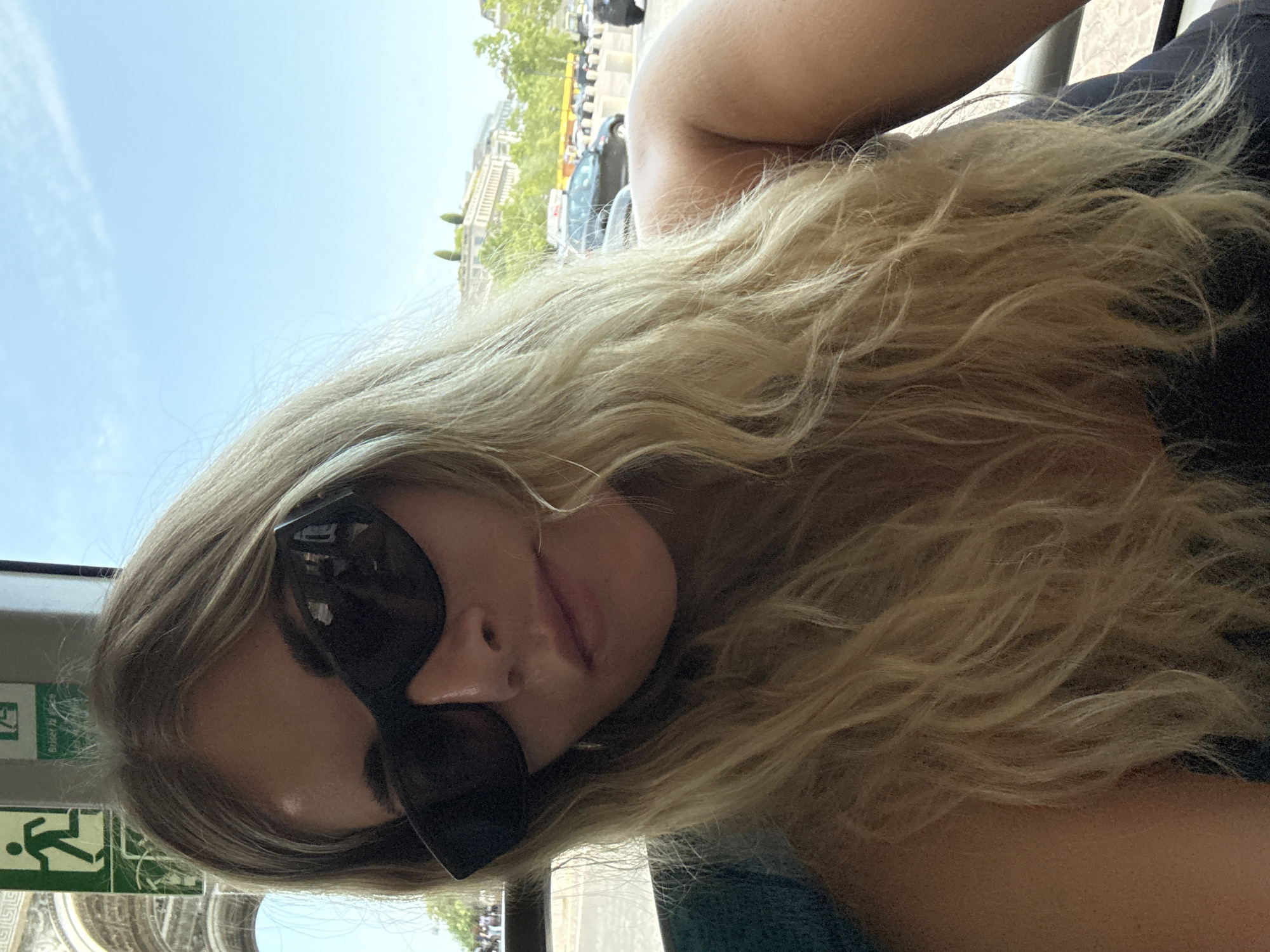 The media file shows a close-up, low-angle, sideways shot of a young woman with blonde, wavy hair and dark sunglasses. She is wearing a dark top, and her face is partially obscured by her hair and sunglasses. Her lips are slightly parted, and she appears to be looking down.
In the background, through a window or opening, there are hints of a city scene. Buildings with ornate architecture, trees, and vehicles can be seen. A green and white sign with an icon of a person running and text is visible on the left side of the frame, suggesting an emergency exit or evacuation route. The lighting suggests it is daytime, with bright, natural light. The overall impression is of someone taking a selfie while traveling or sightseeing in an urban environment, possibly Paris, given the implied architectural style and the presence of the emergency exit sign which is common in public transport.