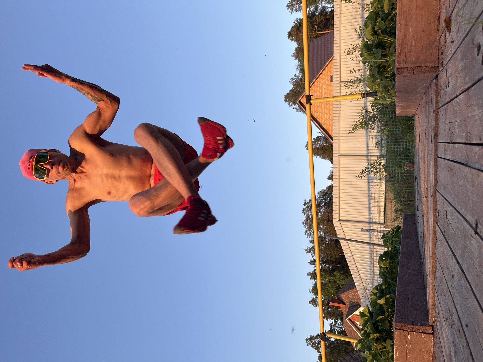 A shirtless man is mid-air, appearing to perform a flip, against a blue sky backdrop. He wears a pink cap, yellow-tinted sunglasses, red shorts, and red sneakers. Tattoos are visible on his left arm. In the background, there's a wooden structure, possibly a platform or deck, adjacent to a fence and a house. The location is Chaikovsky, Russia. Vegetation is growing near the fence. A yellow metal pole is visible along the fence line.