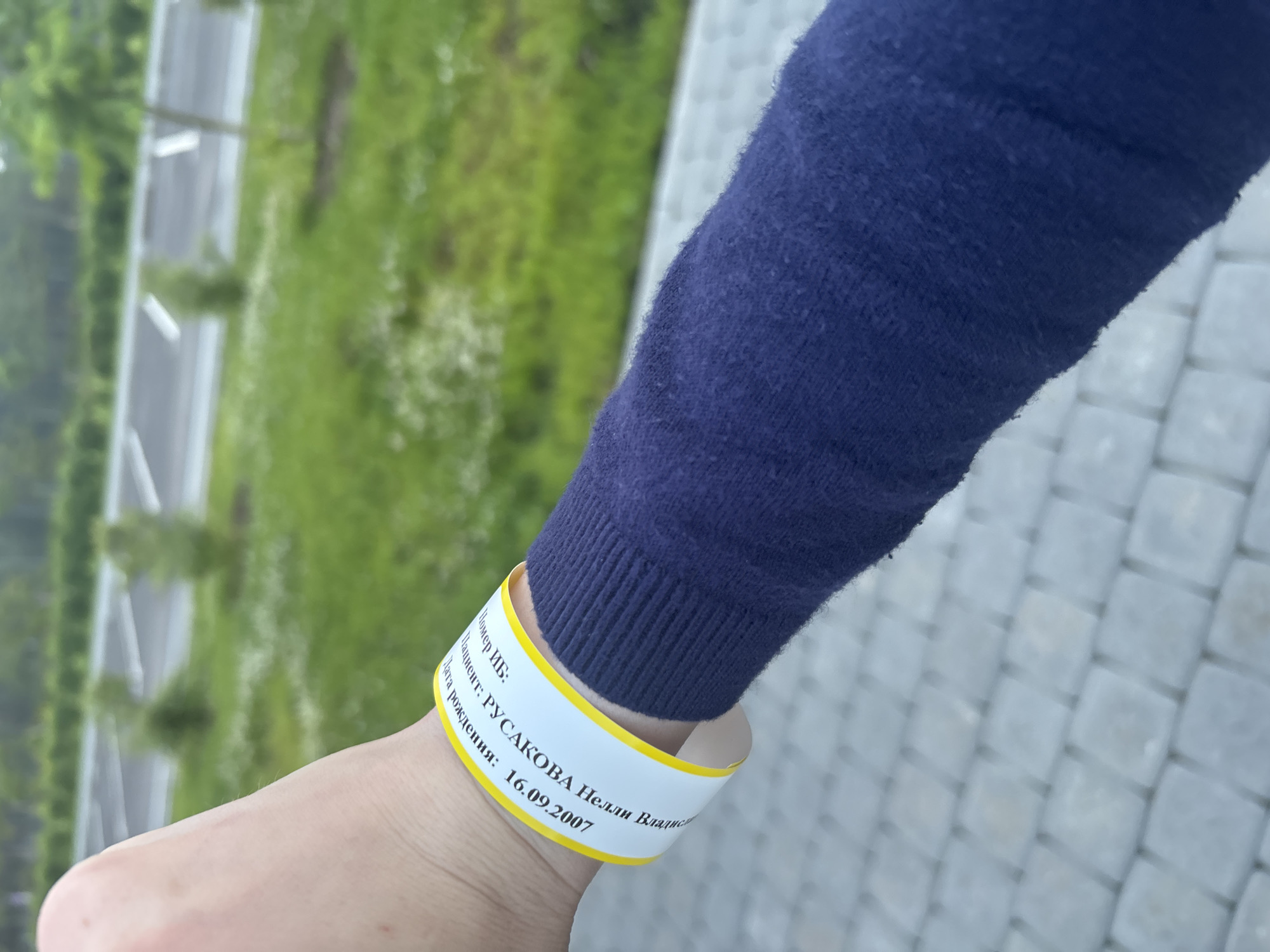 The image shows a close-up of a person's left arm and wrist, wearing a dark blue sweater and a white wristband with yellow trim. The wristband has text printed on it in black, which appears to be in Russian. The visible text on the wristband reads: "Дата рождения: 16.09.2007" (Date of birth: 16.09.2007), "Пациент: РУСАКОВА И.В." (Patient: RUSAKOVA I.V.), and "Номер ИВ:" (IV number:). The background is out of focus, showing a green grassy area with some white flowers and what appears to be a paved area with parking lines. The scene is likely outdoors, possibly in a park or recreational area. While the image does not explicitly show the city of Solnechnoye, Russia, the presence of Russian text on the wristband suggests a possible connection to this location.