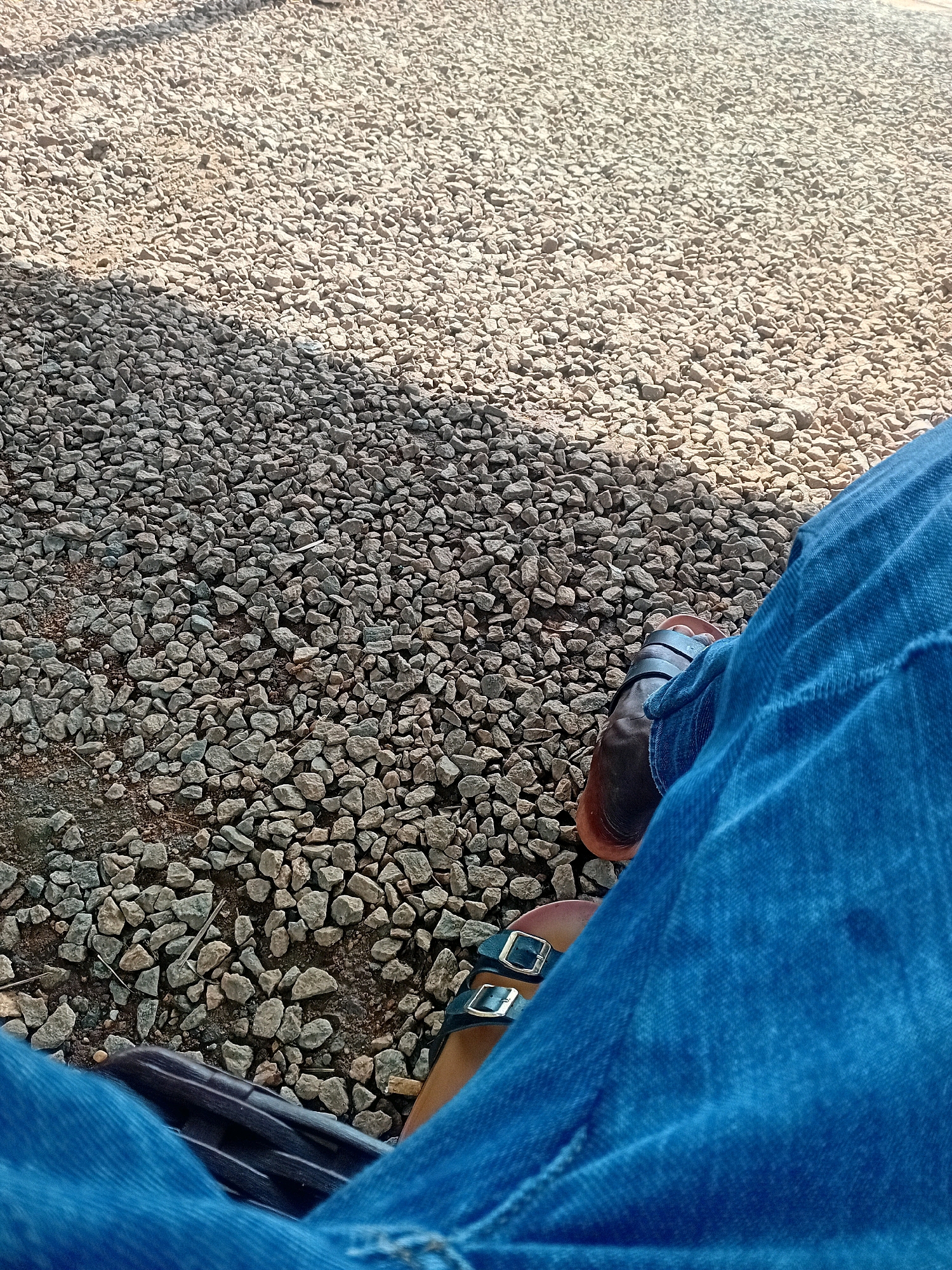 The image shows a person's legs and feet, wearing blue jeans and sandals, sitting on a surface of small grey and beige gravel. The gravel extends throughout the frame, with some areas appearing lighter due to sunlight. A shadow is cast across the gravel from the upper left of the image, suggesting the presence of an unseen object or structure. The person's right foot is visible with a brown sandal, and the left foot has a dark green sandal with two gold buckles. The jeans are denim and appear to be slightly worn. The scene is outdoors and appears to be a public space, possibly a pathway or seating area, in Kira, Uganda.