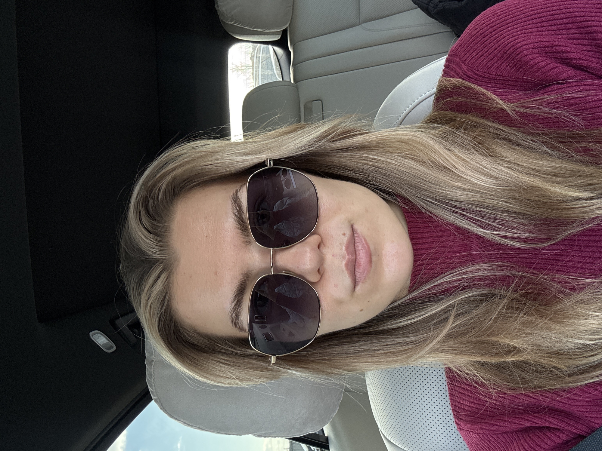 A person, likely female, is featured in a close-up, eye-level shot taken inside a car during the daytime. She has long, wavy blonde and light brown hair that flows down her shoulders and chest. She is wearing large, aviator-style sunglasses with gold frames, which cover her eyes, reflecting parts of the car's interior and the outside environment. Her lips are slightly parted. She is dressed in a burgundy-colored, ribbed knit sweater or top.
The setting is a car interior with light-colored (possibly cream or white) leather seats, including a headrest and back seats that are visible. The car's ceiling is dark, and a light fixture can be seen. Bright daylight illuminates the scene, streaming in through the car windows. Through a side window, trees and a clear blue sky with some clouds are faintly visible. The overall impression is a casual, relaxed moment while traveling in a car. No visible text is present in the image.