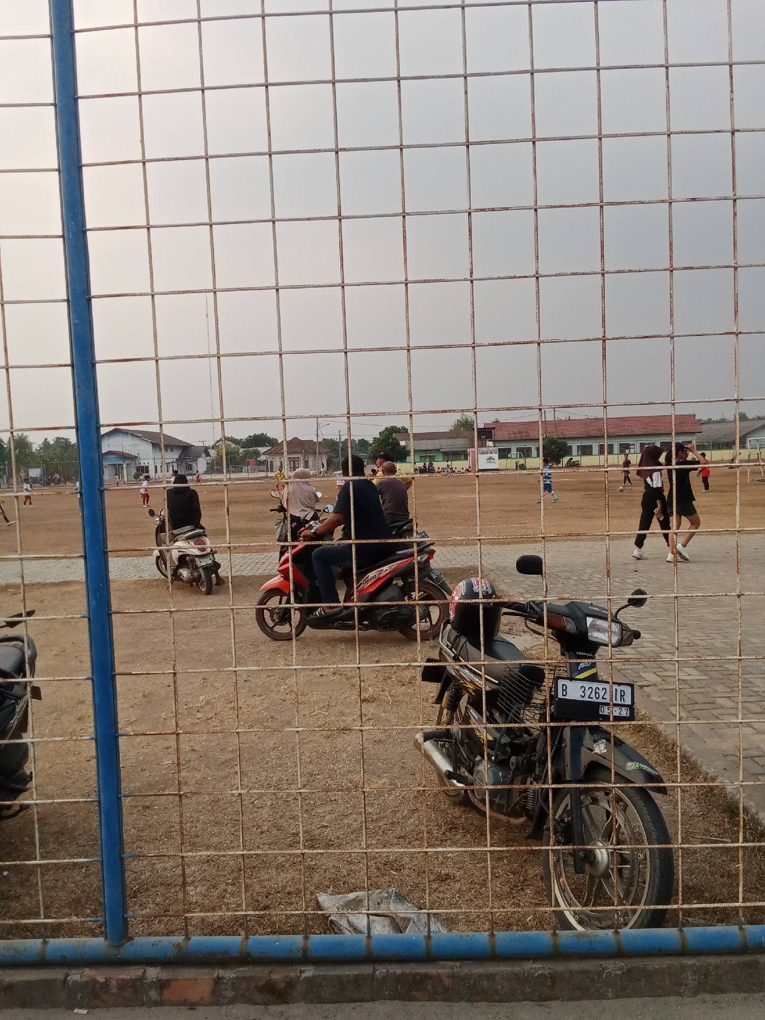 The image shows a view of a dirt field, likely a sports field, through a wire fence. In the foreground, several motorcycles are parked. One black motorcycle with a license plate reading "B 3262 IR" is prominent. Behind the motorcycles, on the field, several people are visible, some engaged in what appears to be a soccer game. Buildings and trees can be seen in the background, suggesting an outdoor location in a developed area. The sky is overcast. The overall impression is a casual scene at a community sports ground. The location is identified as Toto Katon, Indonesia.