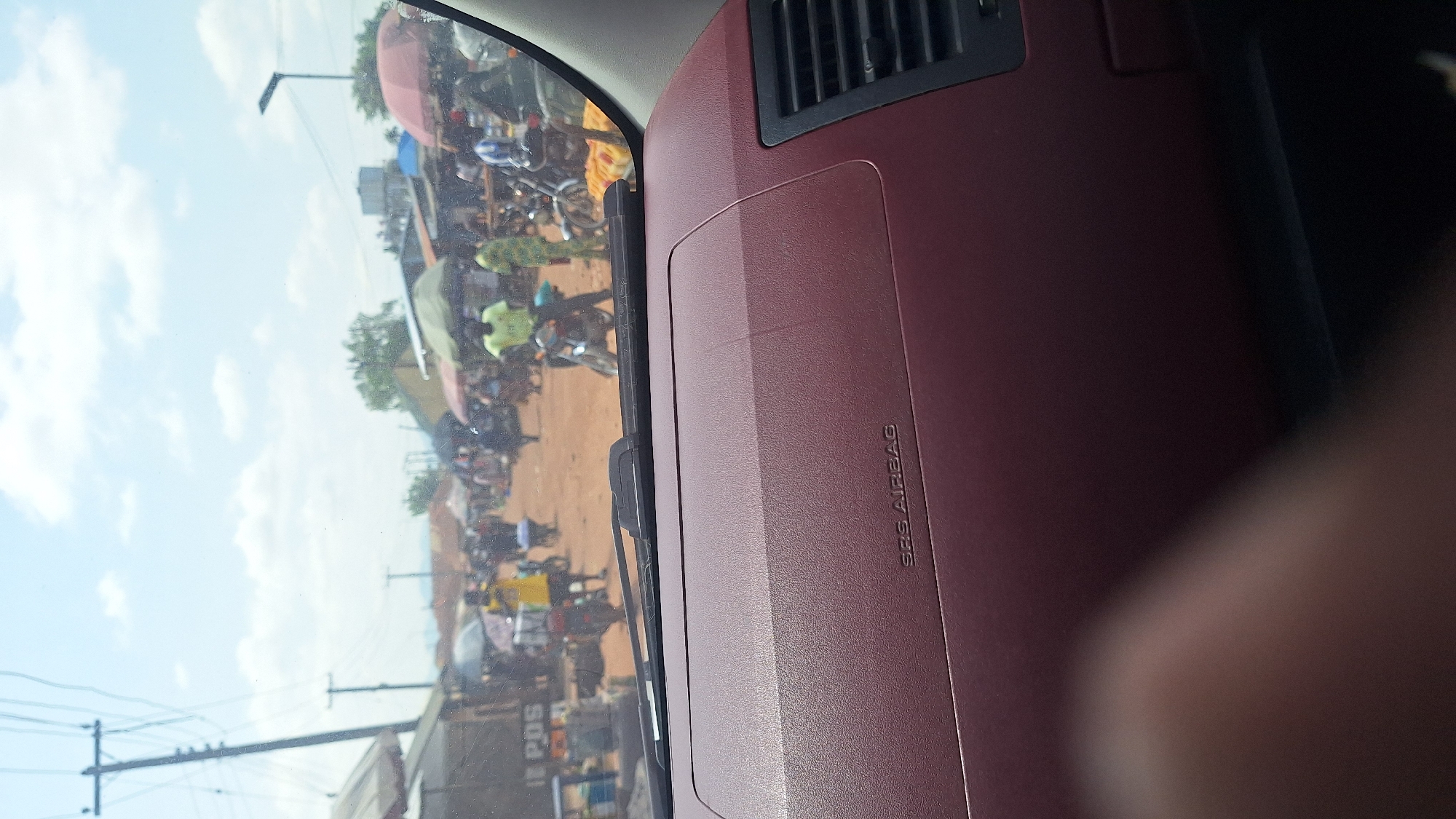 The image shows a street scene, likely a market, in Jalingo, Nigeria. The foreground displays a portion of a car's dashboard, with the words "SRS AIRBAG" visible on the maroon surface. The view through the windshield reveals a bustling environment with numerous people and motorcycles. In the background, stalls and shops line the dirt road, some covered by umbrellas and awnings. Several individuals are seen on motorcycles, and others are walking around, suggesting active commerce and transportation. The sky is partially cloudy.