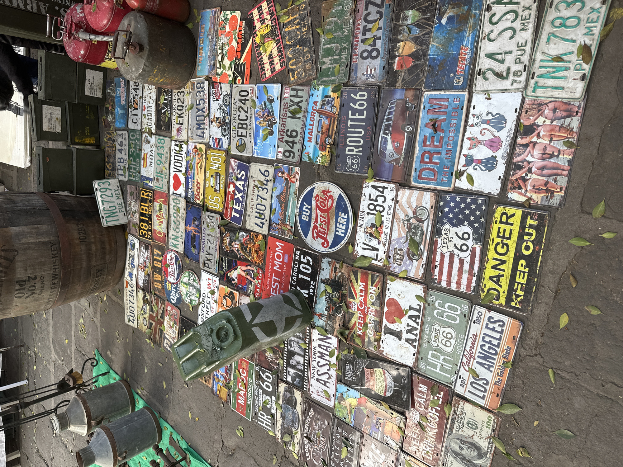 The image shows a street scene in Puebla, Mexico, displaying a collection of vintage and replica license plates arranged on the ground for sale. The license plates are from various U.S. states and Mexico, featuring diverse themes such as Route 66, state slogans, and vintage advertising. Other objects in the scene include a wooden barrel, metal containers, and a green military-style canister. The ground is paved, with scattered leaves. No people are fully visible, although a partial figure is present on the left. The overall impression is a cluttered marketplace setting.