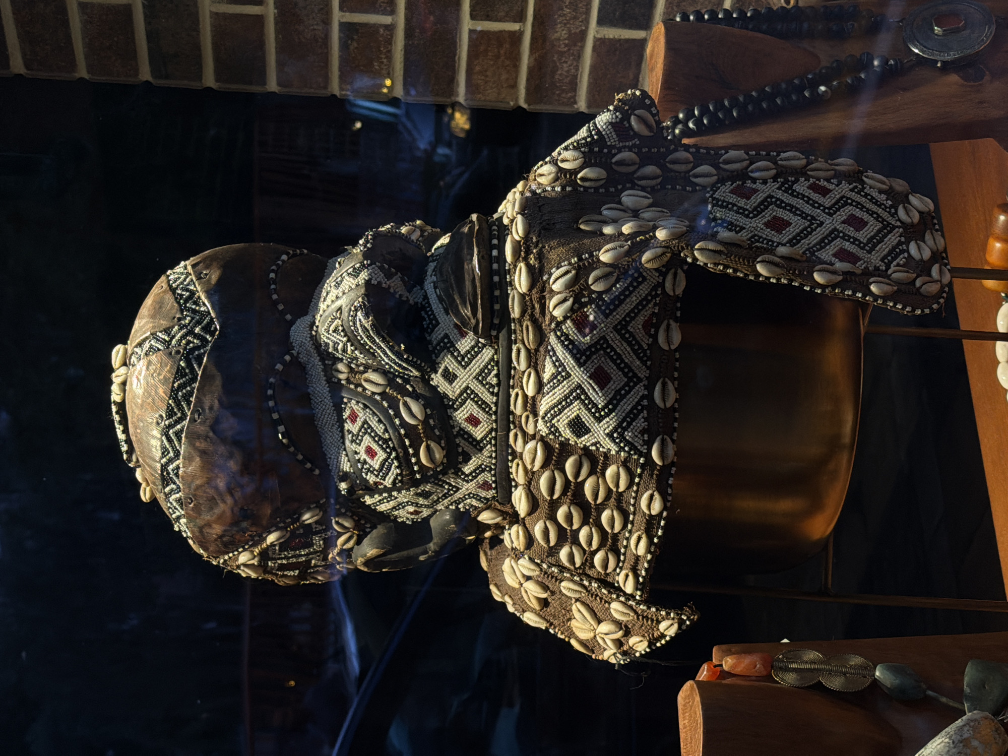 The image shows a ceremonial headdress displayed behind glass, likely in a museum or gallery. The headdress features intricate beadwork and cowrie shell embellishments. The beadwork forms geometric patterns with red and black accents. It's placed on top of a metallic gold colored bowl on a wooden shelf. Another shelf below contains what appears to be stone artifacts and necklaces. In the background, brickwork is visible above the reflection in the glass. The location is the Museum of African Art in Belgrade, Serbia.