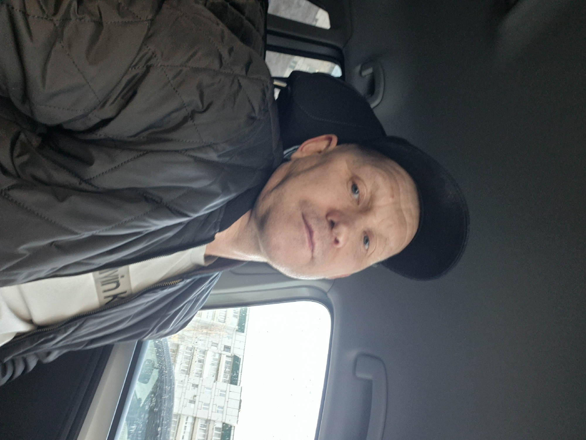 An adult male is depicted in a close-up selfie taken from inside a vehicle, looking directly at the camera. He is wearing a black baseball cap, a black quilted bomber jacket, and a white t-shirt with partial "Calvin Klein" branding visible. The man has fair skin and blue eyes. He is seated in what appears to be the front passenger seat, with a dark headrest behind him. Through the vehicle's side window on the right, a residential urban environment in Chaikovsky, Russia, is visible. The background features several light-colored, multi-story apartment buildings with numerous windows and balconies, some exhibiting green roof or awning details. The sky is overcast, and the vehicle window shows evidence of light precipitation. A dark vehicle is partially obscured in the distant background.