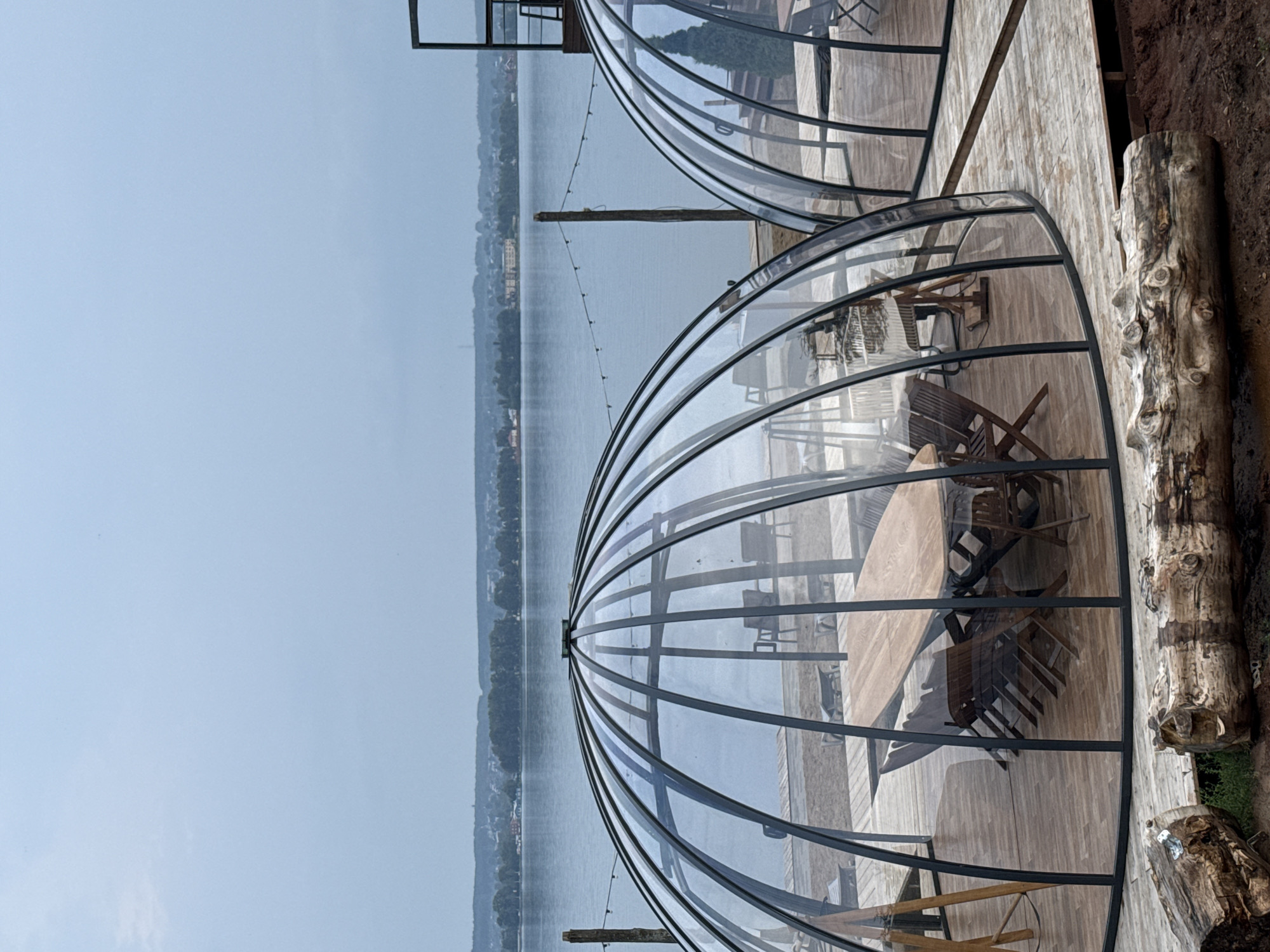 The image depicts a waterfront scene with two geodesic dome structures made of clear plastic and dark metal framing. These domes are situated on a wooden deck overlooking a body of water, likely a lake or river, with a tree-lined shore and buildings in the distance. The sky is overcast, suggesting a cool or possibly humid day. Inside the foreground dome, a large wooden table and several wooden chairs are visible. The overall atmosphere appears calm and serene, with the natural landscape providing a peaceful backdrop. The domes seem to be designed for outdoor dining or seating, offering shelter while maintaining a connection to the surroundings. There are no people visible in the image, and no visible text. The presence of a thick log on the right side of the frame adds a rustic element to the setting.
