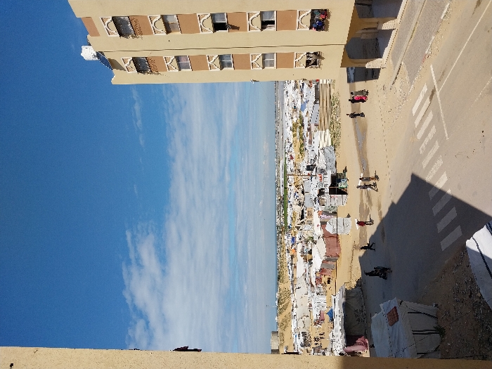 The image depicts an elevated view of a scene in Khan Yunis, Palestinian Territory, under a bright blue sky with scattered white clouds. In the immediate foreground, a paved road with a white-striped pedestrian crossing is visible, partially covered by a dark shadow originating from the viewpoint's building. To the right, a multi-story apartment building, light beige with brown decorative window frames and balconies, features multiple windows (some open), a white water tank, and solar panels on its roof. The left edge of the frame shows a partial, plain beige wall, indicating the vantage point.
The midground and background are dominated by a vast displacement camp, characterized by numerous densely packed makeshift shelters and tents constructed from various materials, primarily white, grey, and brown tarpaulins and fabrics. Several individuals are present: a solitary figure near a shelter on the left, a group of three people on the dirt path carrying a long, light-colored object, and another group of three individuals (one in red) walking further right. Additional smaller figures are scattered across the camp area.
Beyond the camp, the landscape includes sandy dunes and patches of green vegetation. The distant horizon reveals a large body of water, identifiable as the Mediterranean Sea, with faint industrial or urban structures visible along the coastline. A prominent cylindrical structure stands in the far left background.