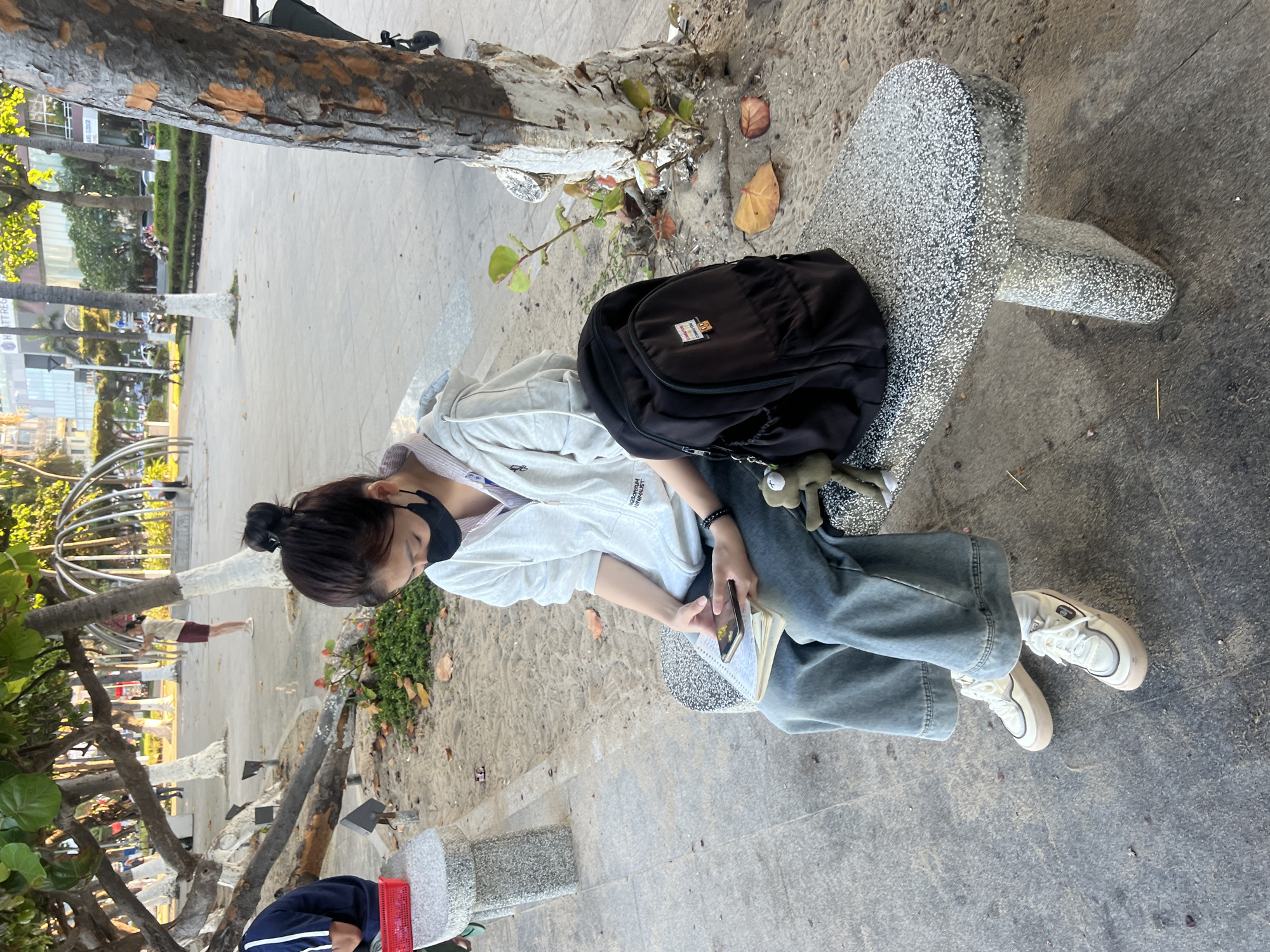 The image displays a young female seated on a speckled gray stone bench outdoors in Nha Trang, Vietnam. The individual wears a black face mask, a light gray zip-up hoodie over a striped shirt, loose light-wash denim jeans, and white/beige platform sneakers. She is looking down at a black smartphone held in both hands, with a spiral-bound notebook resting on her lap. A black backpack with a green frog-like keychain is positioned on the bench next to her.
The ground surrounding the bench consists of packed sand or dirt, interspersed with fallen dry leaves and some green low-lying foliage. Several trees with exposed roots and partially white-painted trunks are visible throughout the scene. To the left, a segment of a concrete pathway is present. In the background, another individual is partially visible on a separate gray bench. Further back, a paved area transitions into a street with visible vehicle traffic and several multi-story buildings, one featuring the text "HOTT REG" and another "ANG LODGE." The overall lighting indicates natural daylight conditions.