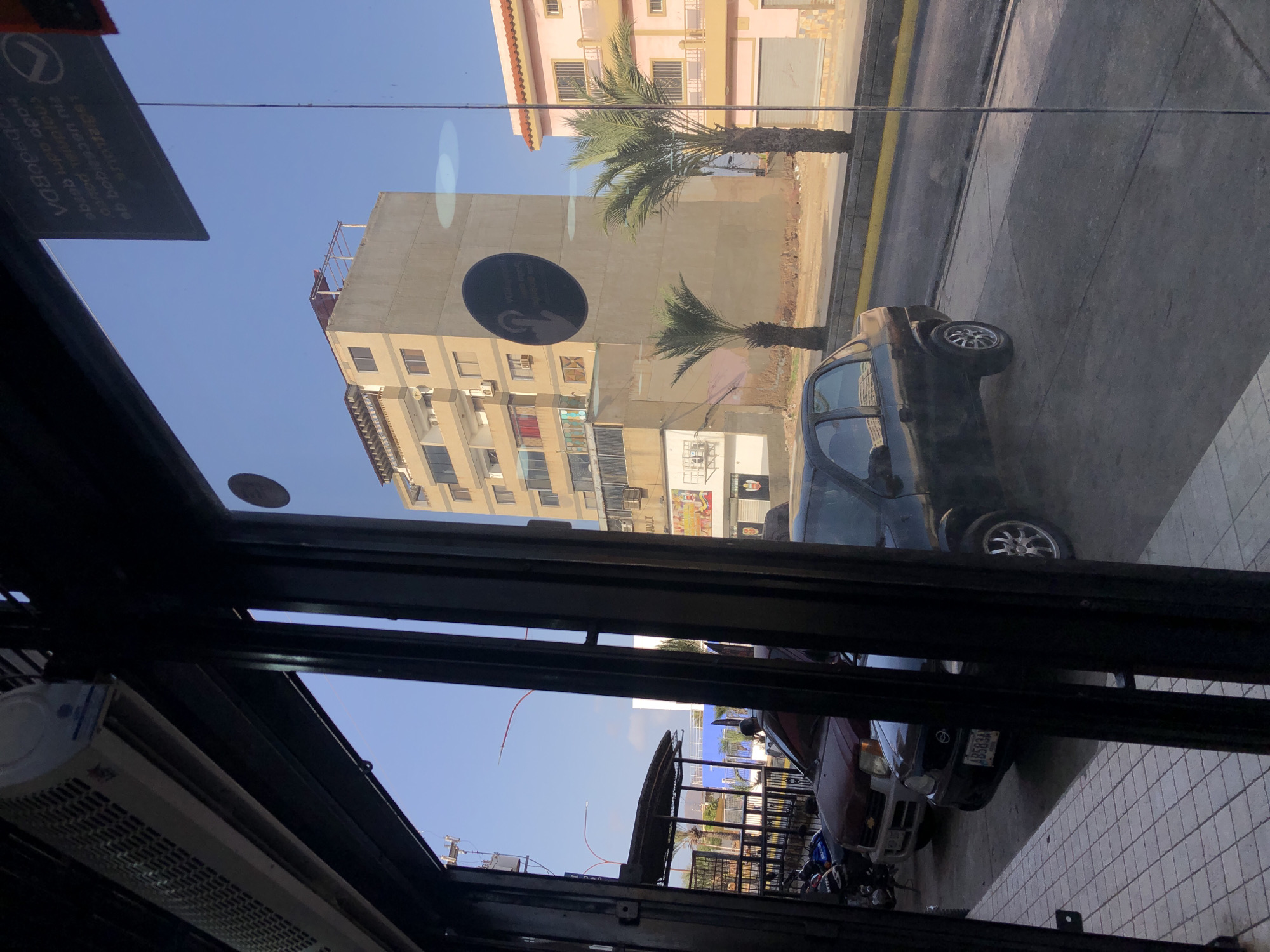 The image depicts an urban scene in Juan Griego, Venezuela, as viewed through a window. Reflected in the window is a dark-colored car parked on a street lined with a sidewalk and buildings of varying heights and architectural styles. The buildings range from multi-story residential structures to commercial establishments, one with visible graffiti. A palm tree is seen along the street. The window frame is visible at the edges of the image, and an air conditioning unit is seen near the bottom left. Below and to the right of the dark-colored car, another car, a light-colored SUV, and a motorbike, can be seen parked on the sidewalk.