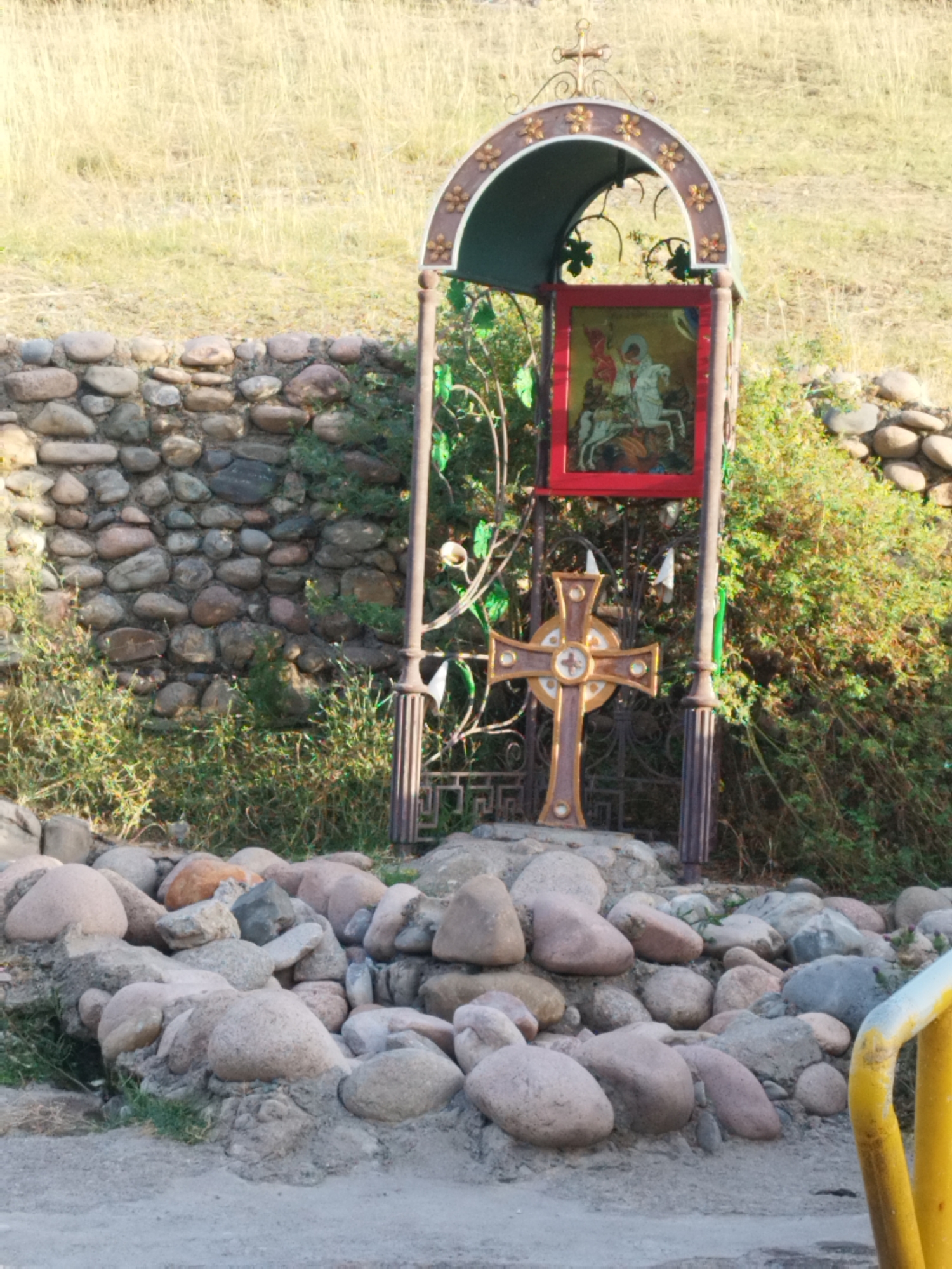This image shows an outdoor scene with a stone wall and dry grass in the background. In the foreground, there is a religious shrine featuring a metal cross with an icon of Saint George slaying the dragon displayed above it. The shrine is decorated with metalwork and stands on a foundation of rocks. The surrounding environment includes more rocks and some vegetation. The scene appears to be located in a rural or natural setting, possibly in or near the village of Koksayaq in Kazakhstan.