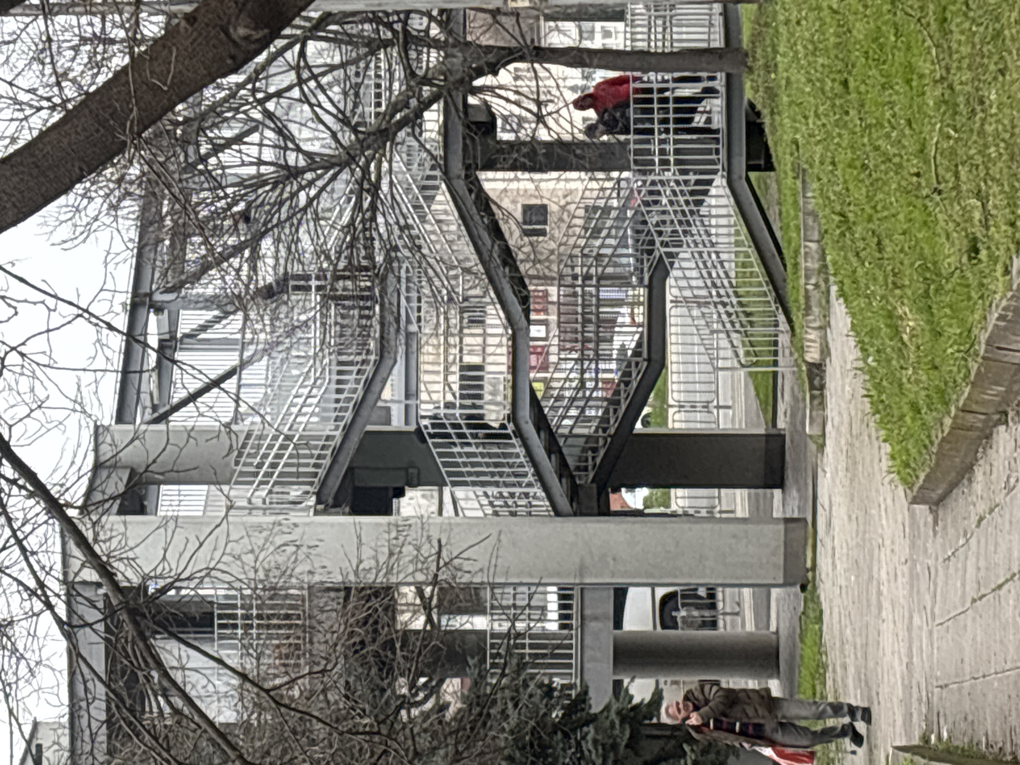 The image shows a building with exposed metal staircases. A person wearing a red jacket is visible on one of the staircases. Below, a person is walking on a sidewalk next to a patch of grass. Bare tree branches are visible in the foreground. The location is likely in Burgas, Bulgaria, as the building appears to be part of a campus or public building with modern architectural elements. The time of year is likely late autumn or early spring, indicated by the bare trees and dormant grass.