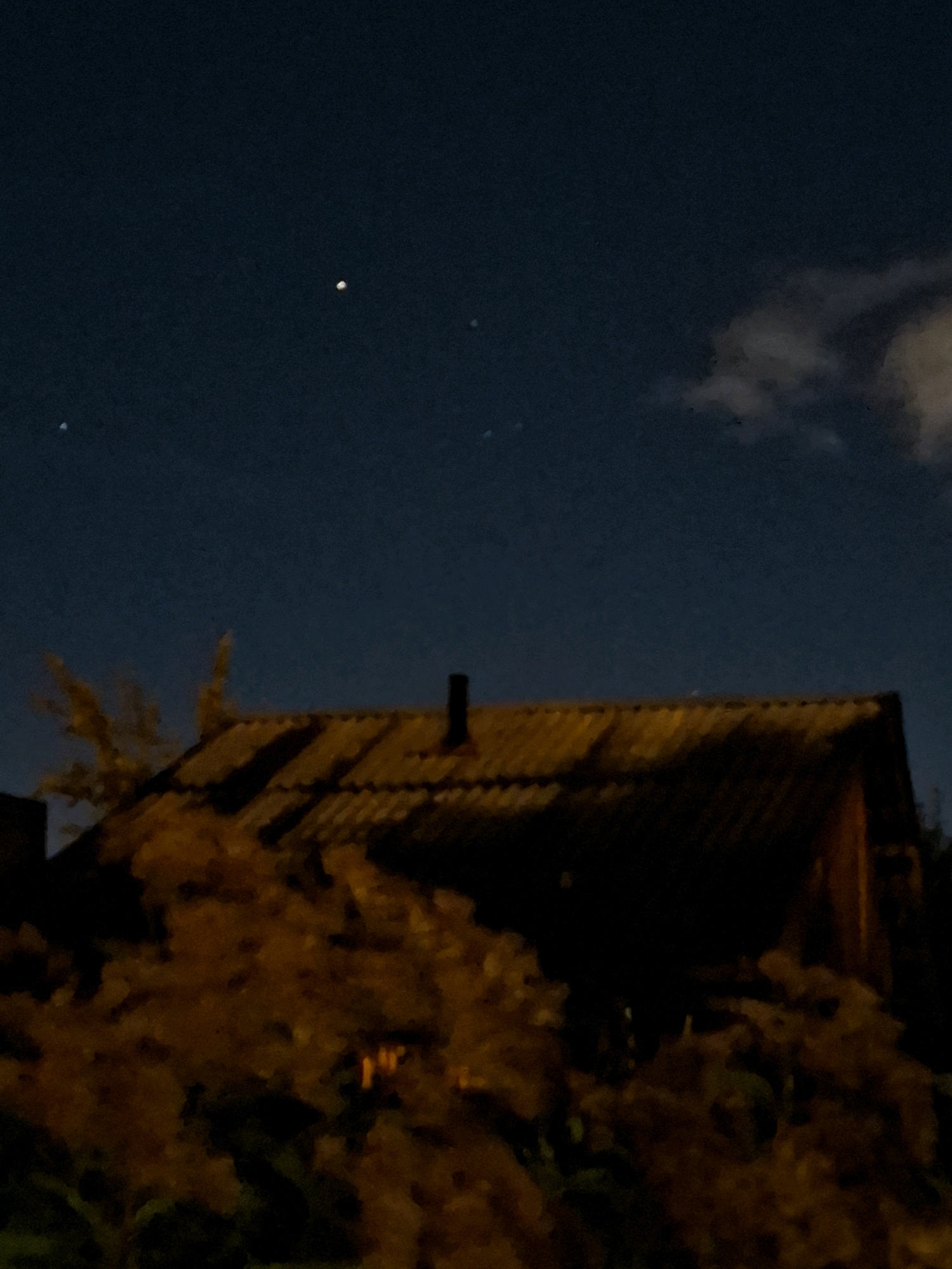 The image shows a nighttime scene with a dark blue sky. Three bright points of light are visible in the sky, one larger and brighter than the other two. A dark, sloped roof of a building, possibly a house, is visible in the foreground, with a chimney. The roof appears to be made of corrugated material. In front of the roof, a mass of dark foliage, likely trees or bushes, is present, illuminated with an orange glow from an unseen light source. The scene is set in the city of Tyumen, Russia.