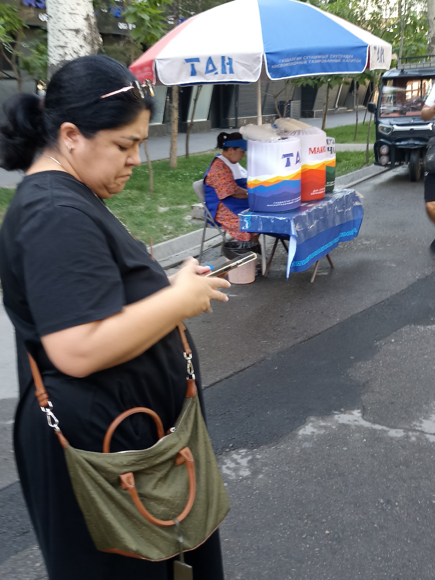 A woman is walking on a street in Bishkek, Kyrgyzstan, holding a smartphone. She is wearing a black dress and carrying a green bag. In the background, there is a vendor selling drinks under an umbrella. A small electric vehicle is parked nearby. The street is paved and there is grass and trees on the side.