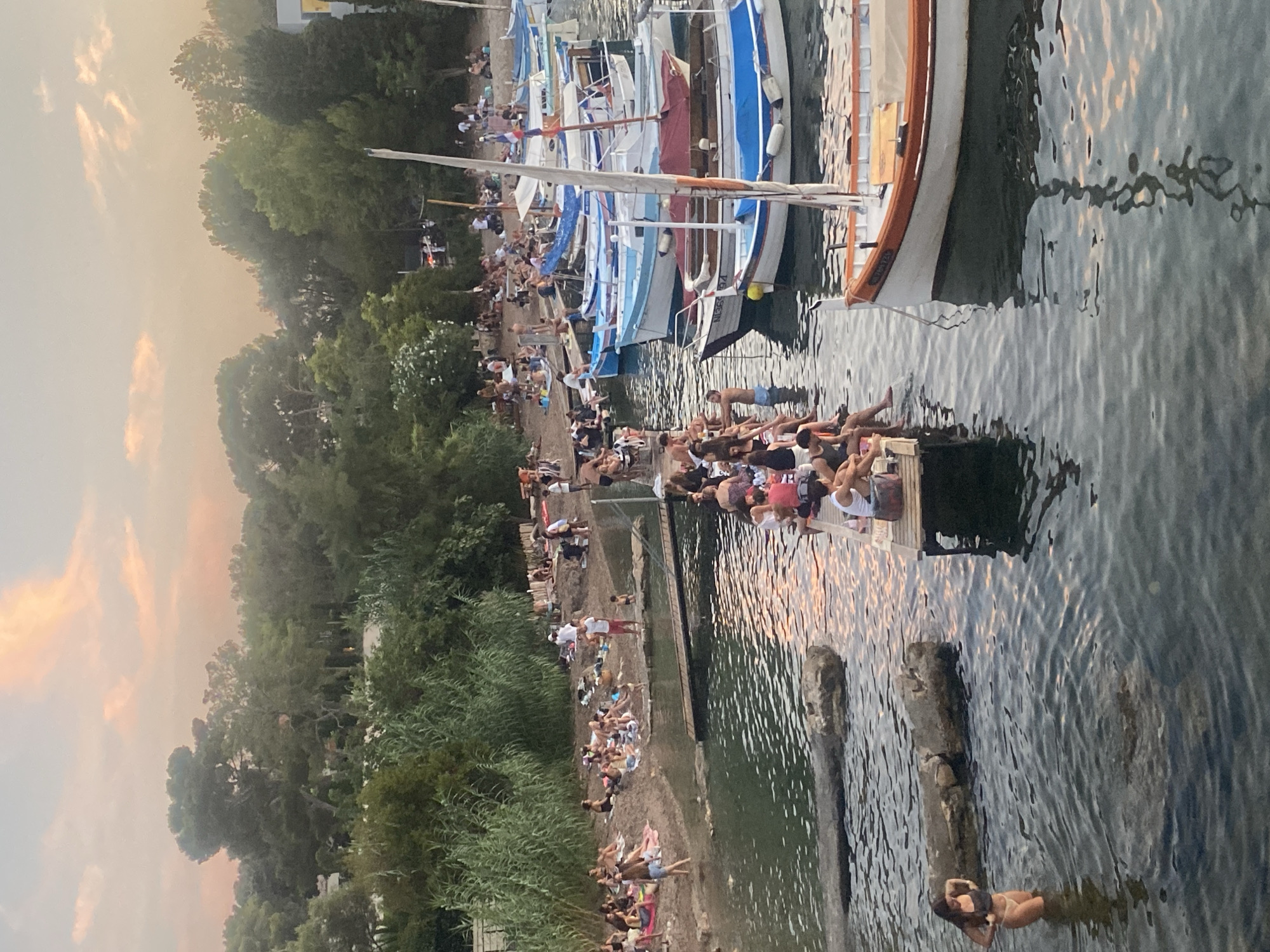 This is an aerial, eye-level, medium shot of the beach and harbor in Antibes, France, during sunset. The scene is bustling with activity, with many people enjoying the warm evening. In the foreground, a woman in a bikini stands in the shallow water, adjusting her swimwear. To her right, a raft-like structure floats in the water, crowded with people sitting and standing. Further back, the sandy beach is populated by numerous individuals, some sunbathing on towels and others standing and mingling. Several boats are moored in the harbor, their reflections shimmering on the water's surface. Lush green trees line the background, and the sky is a soft blend of pink and orange hues, indicating the setting sun.